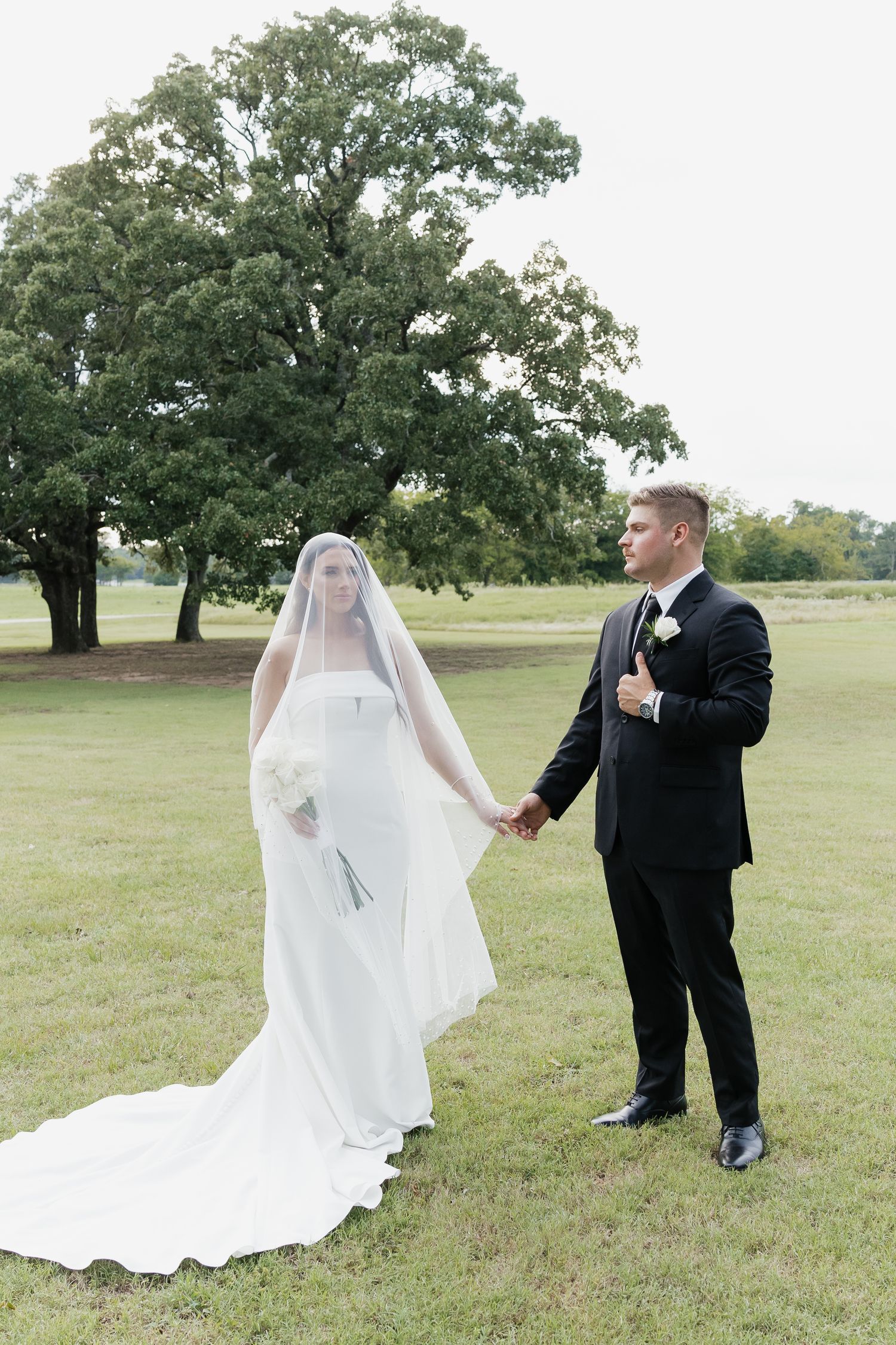 Bride in white gown and veil holds hands with groom in black suit on green lawn with large oak tree.