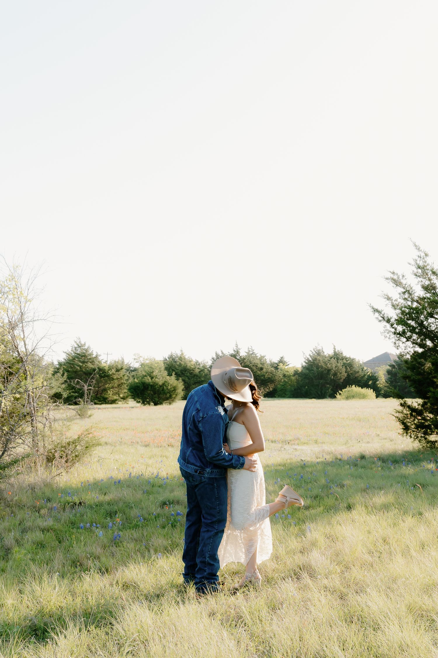 Couple sharing a kiss in open green field surrounded by trees during golden hour.