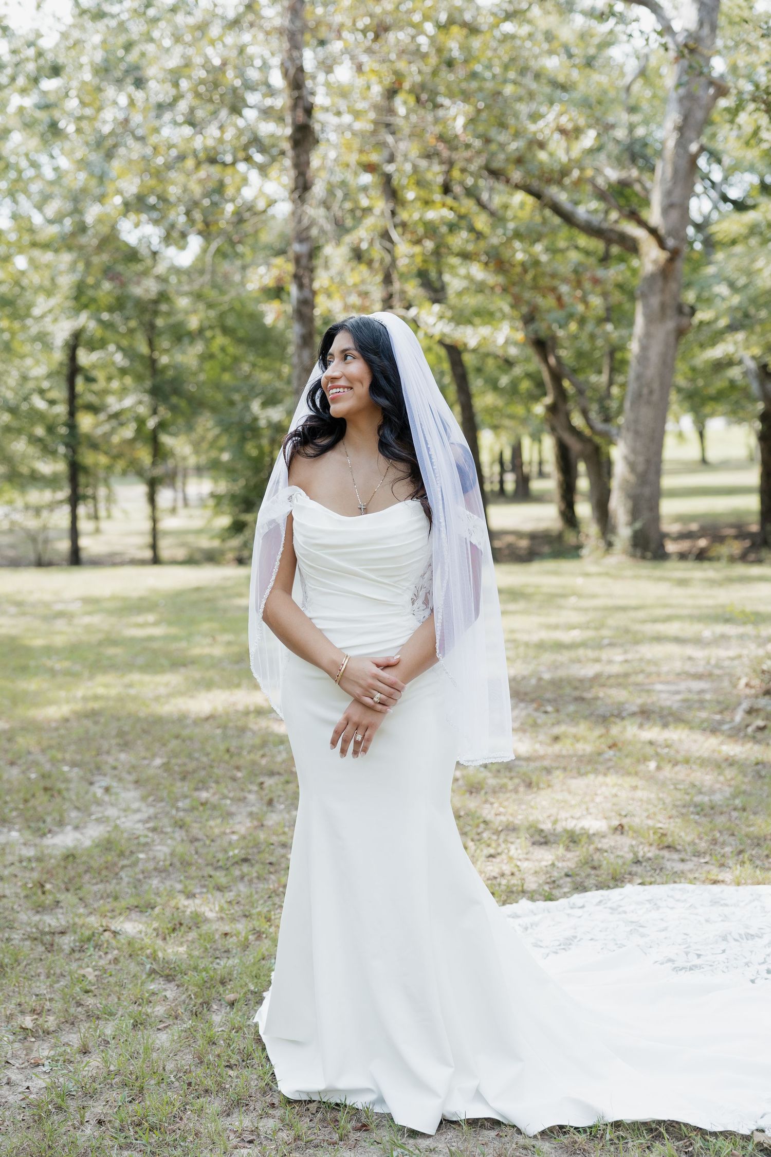 Elegant bride in white mermaid gown and veil smiles while standing among trees outdoors.