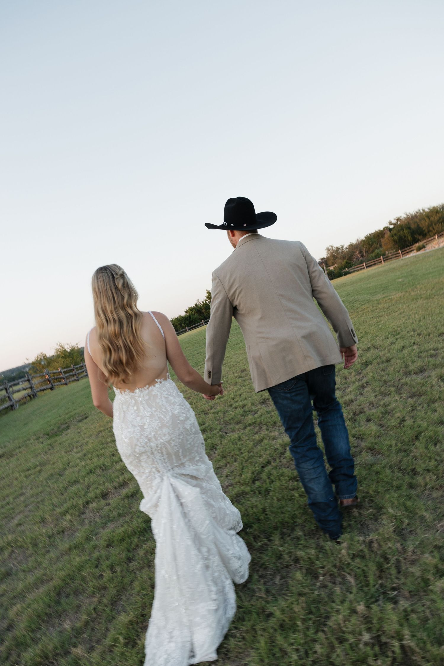 Bride and groom walk hand in hand across green field at sunset, groom in cowboy hat.