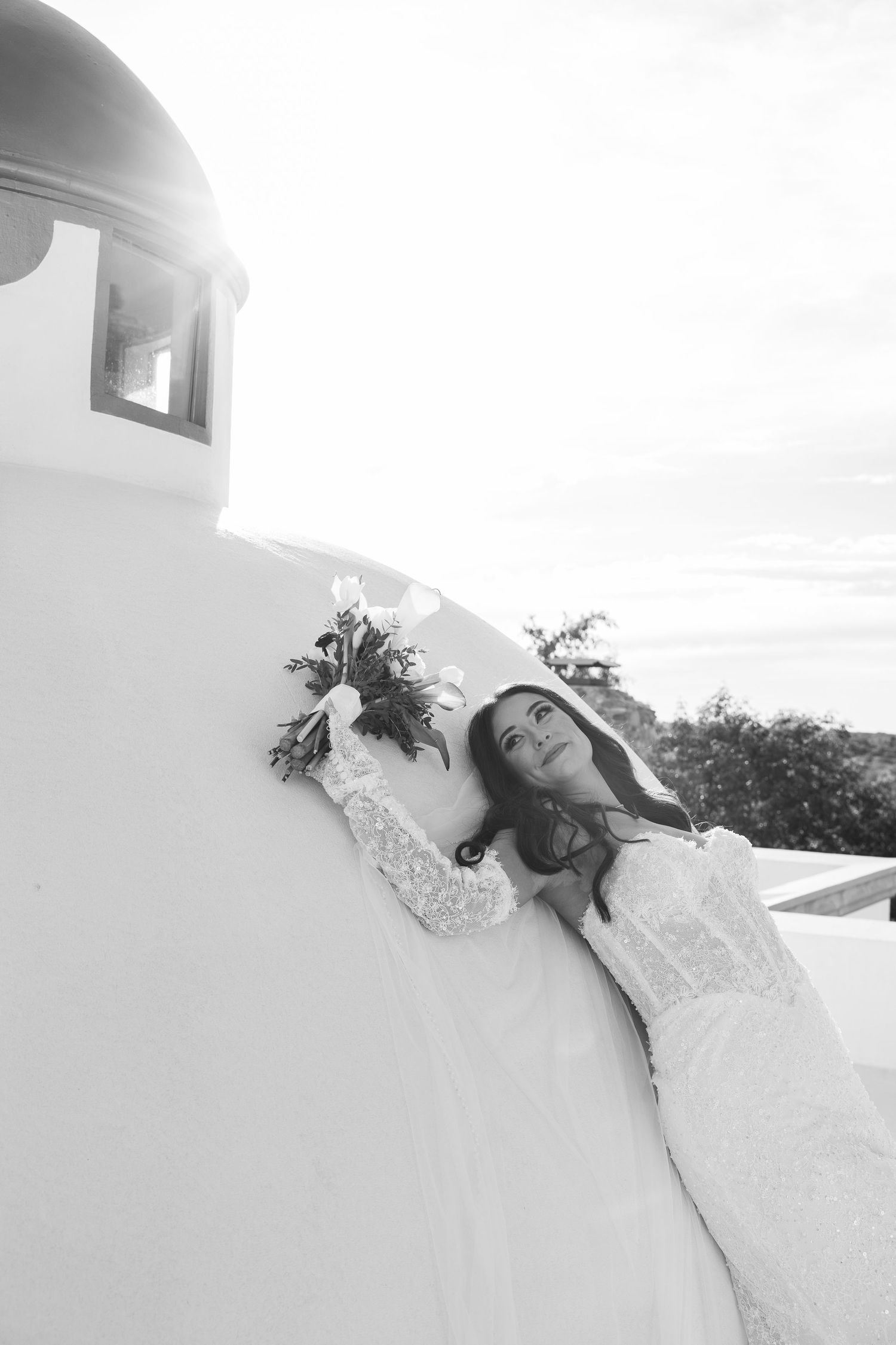 Black and white photo of a bride in lace dress leaning against white Santorini wall holding a floral bouquet.