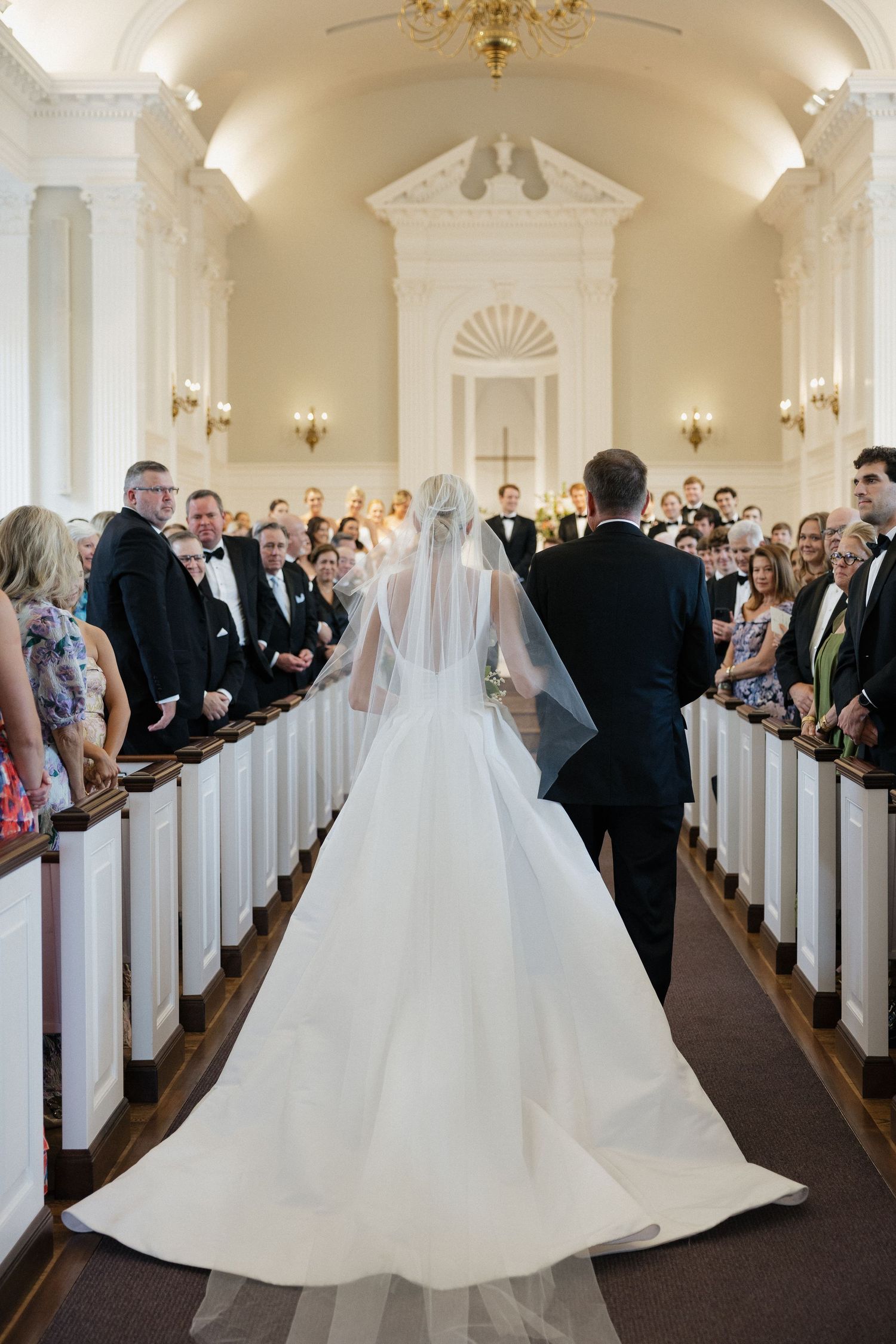 Bride walking down church aisle in elegant white gown with long veil, escorted by father past seated wedding guests.