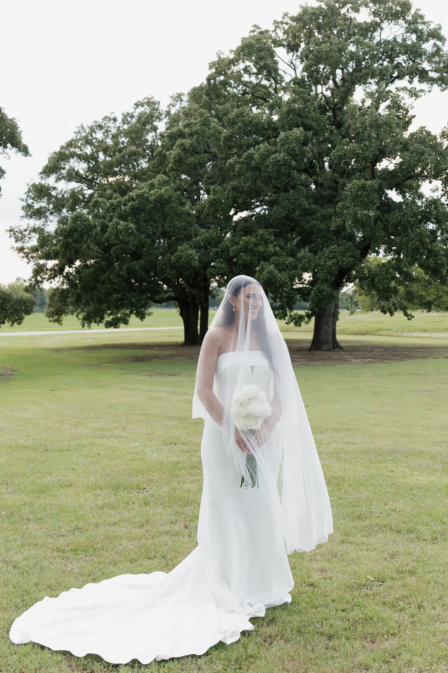Bride in white strapless gown and veil holds bouquet outdoors near large oak trees on green lawn.