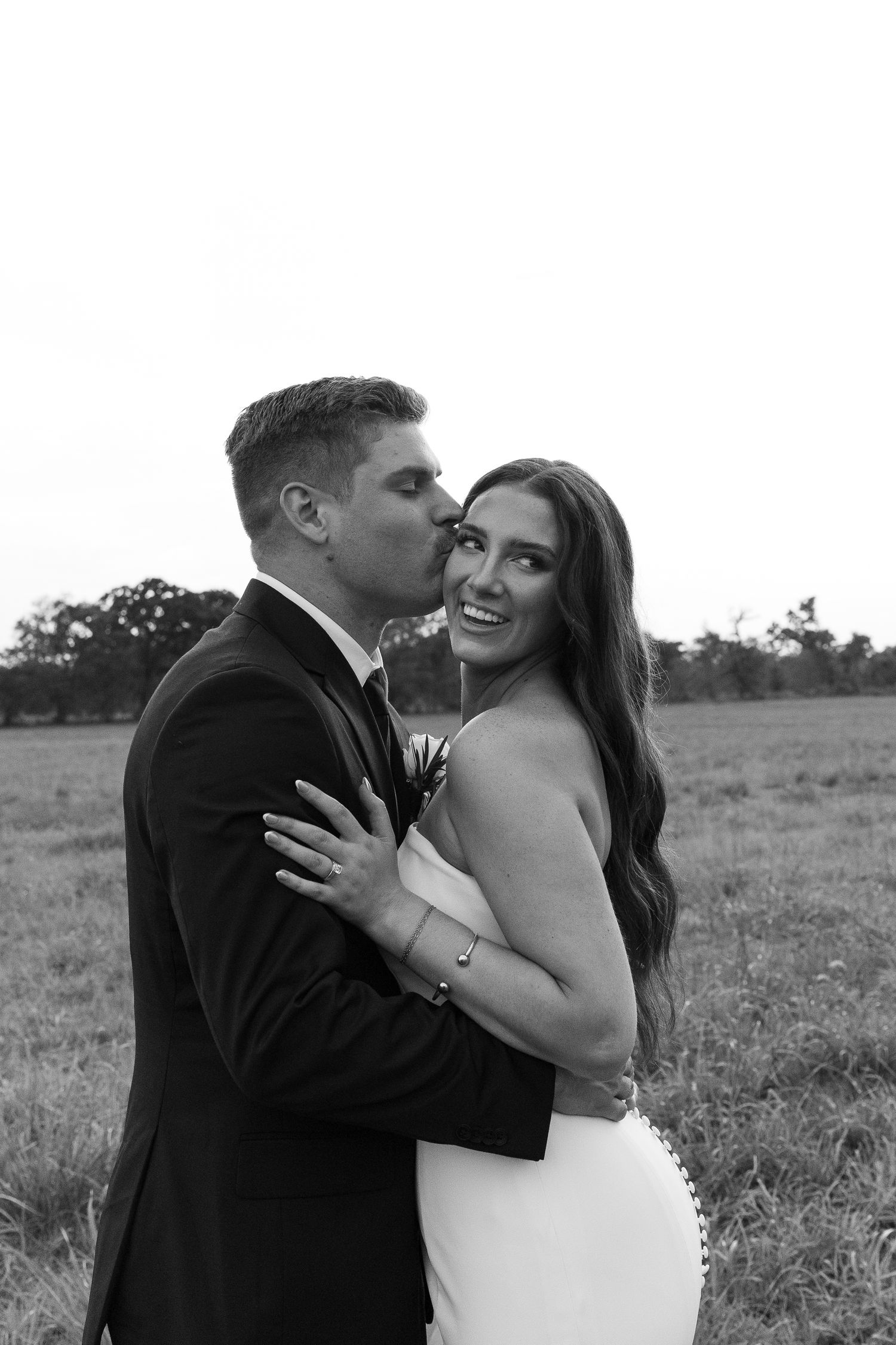 Black and white photo of groom kissing brides cheek as she smiles in open field at sunset.