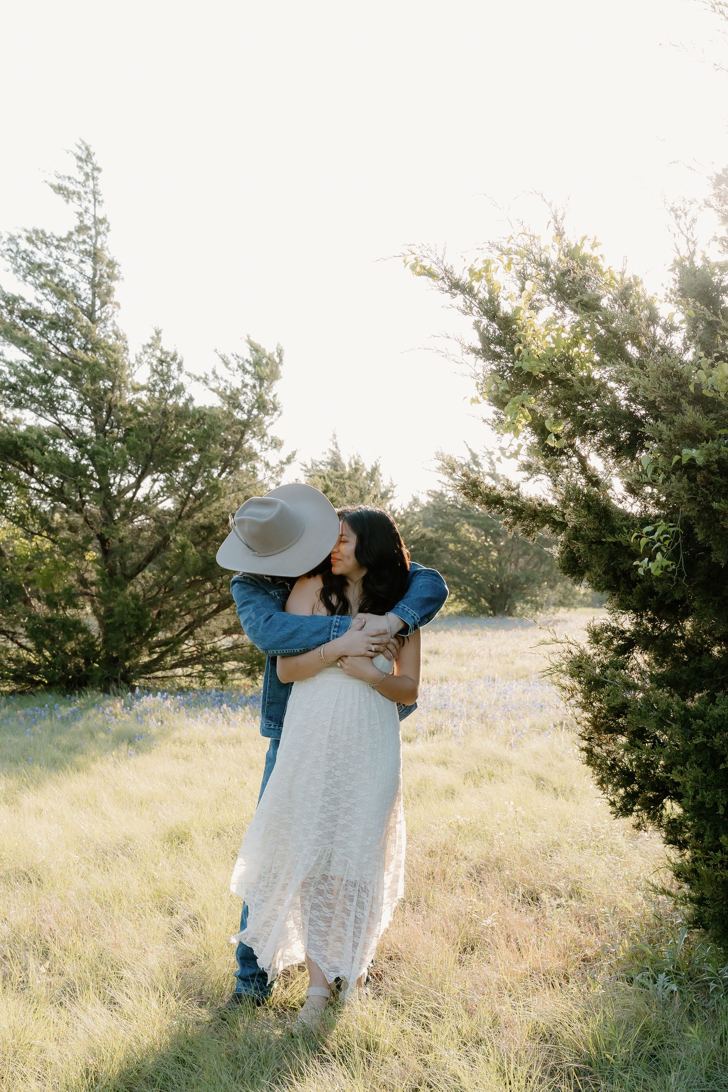 Man in cowboy hat embracing woman in white lace dress in sunlit field with cedar trees.