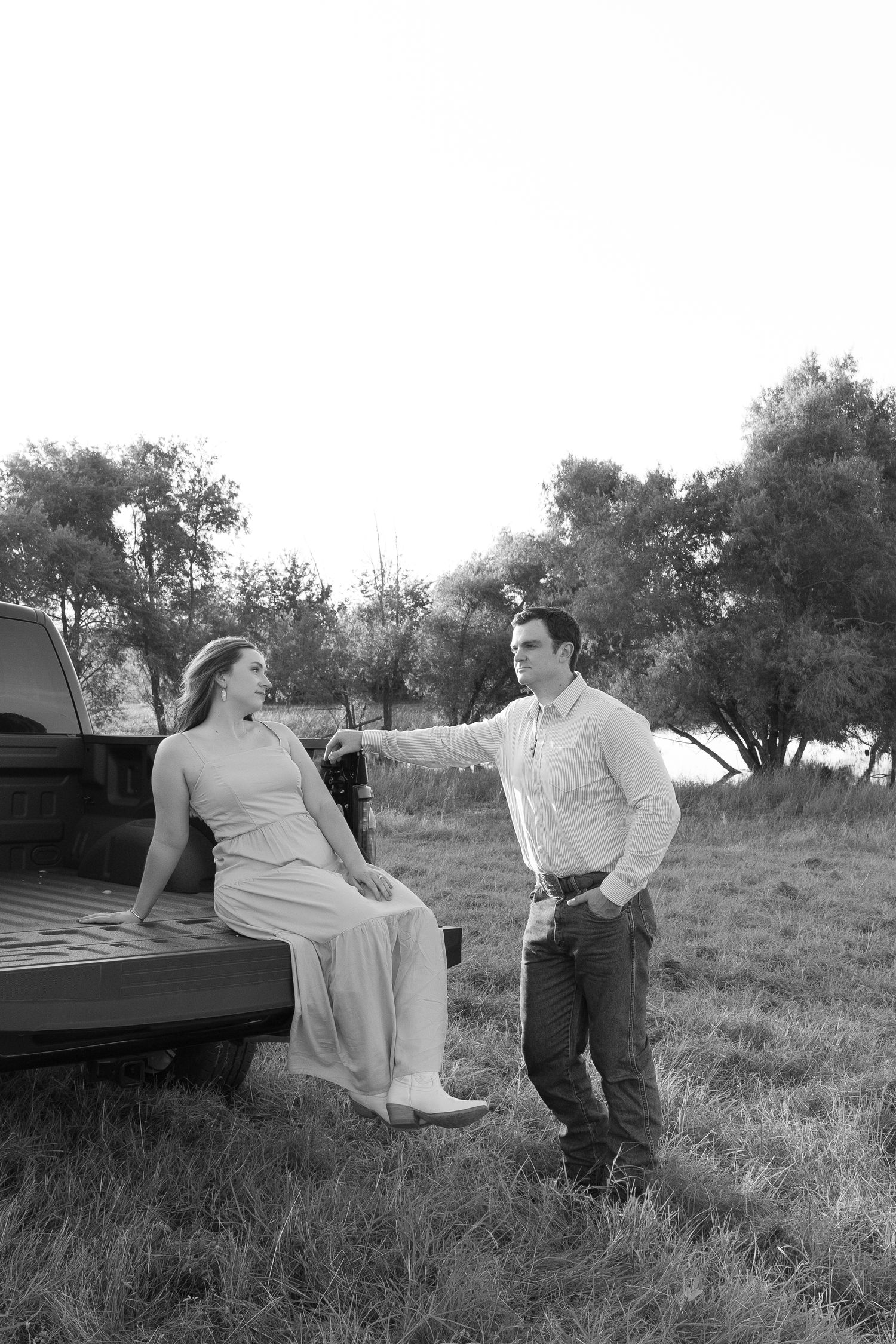 Black and white photo of couple posing on a truck tailgate in an open field at sunset.