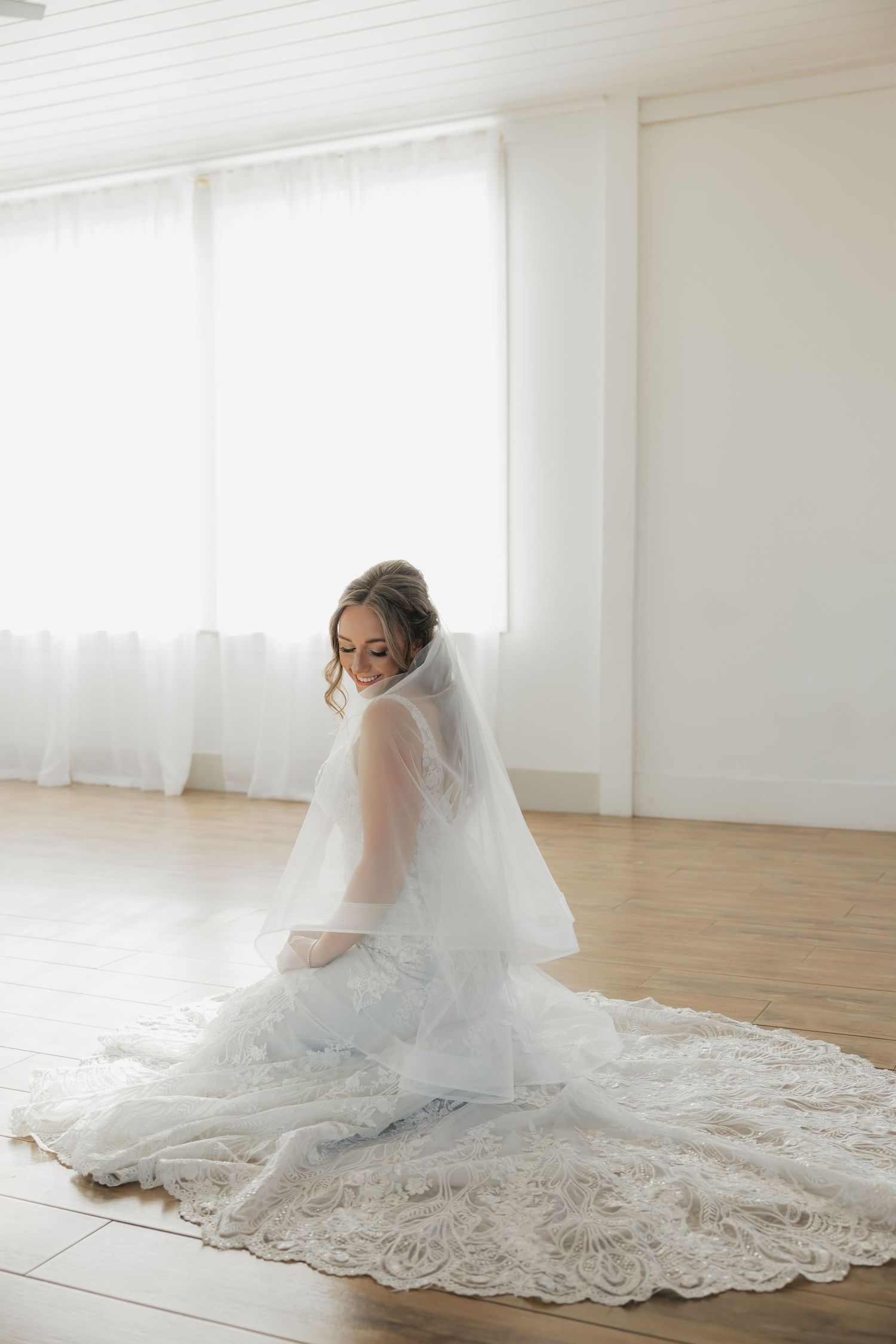 Bride in lace wedding dress and veil sitting gracefully on hardwood floor.