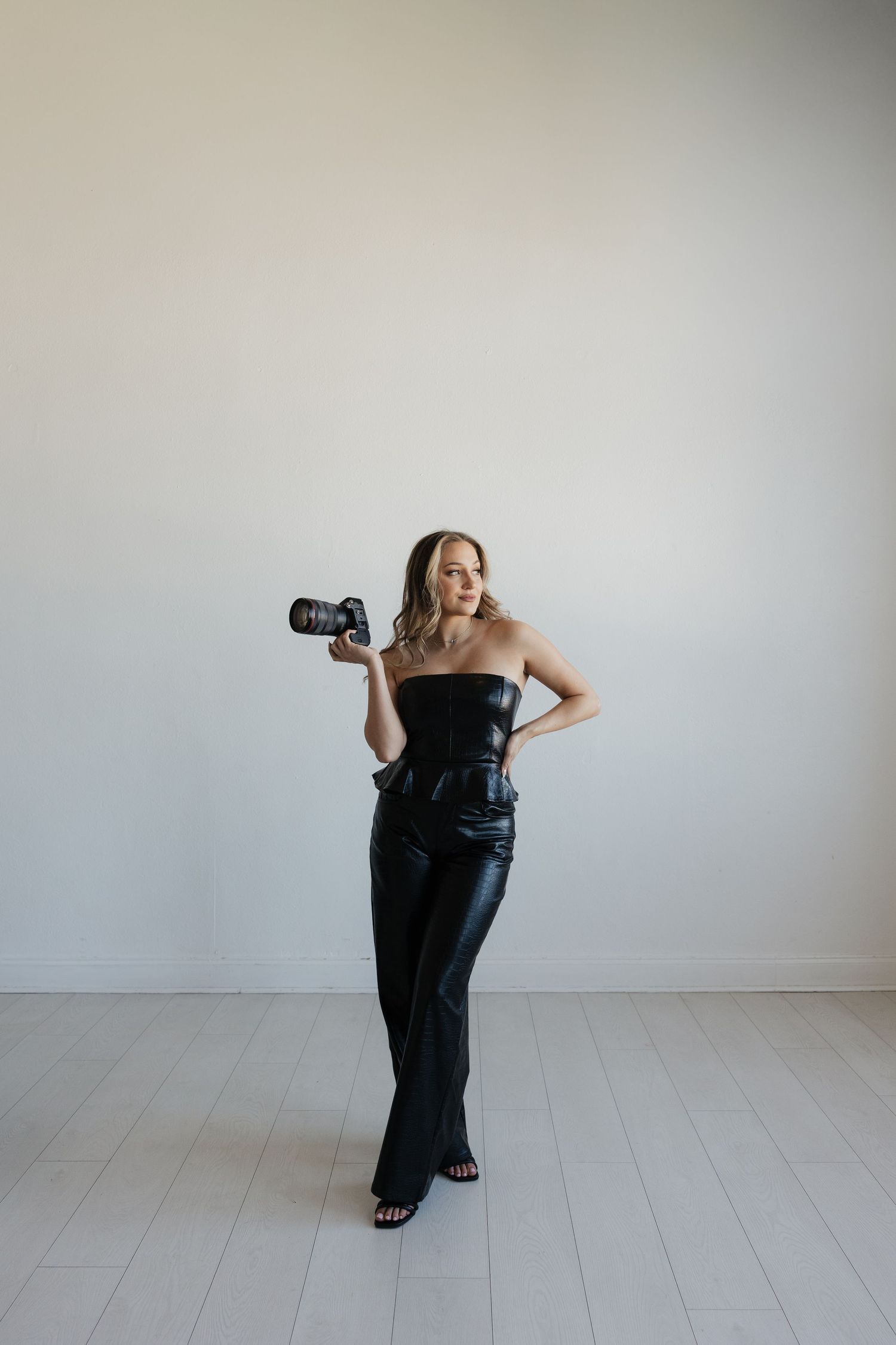 Female photographer in black strapless outfit holding camera against white wall.
