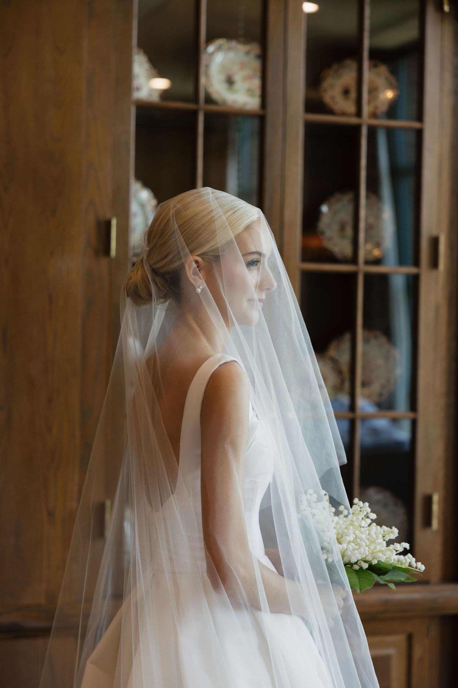 Elegant bride in white gown and layered veil holding lily of the valley bouquet beside wooden cabinet.