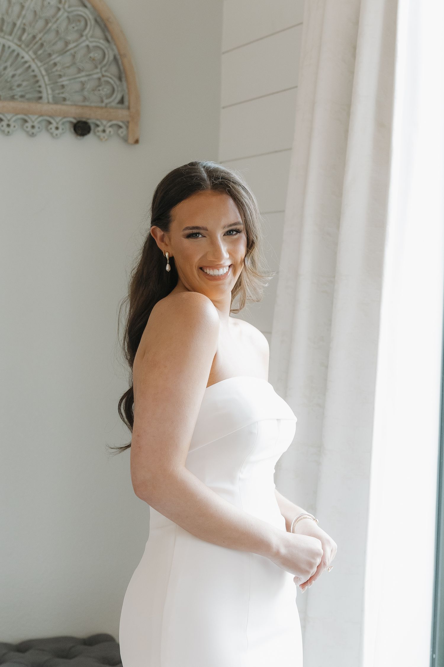 Smiling dark-haired bride in strapless white mermaid gown poses indoors by a bright window.