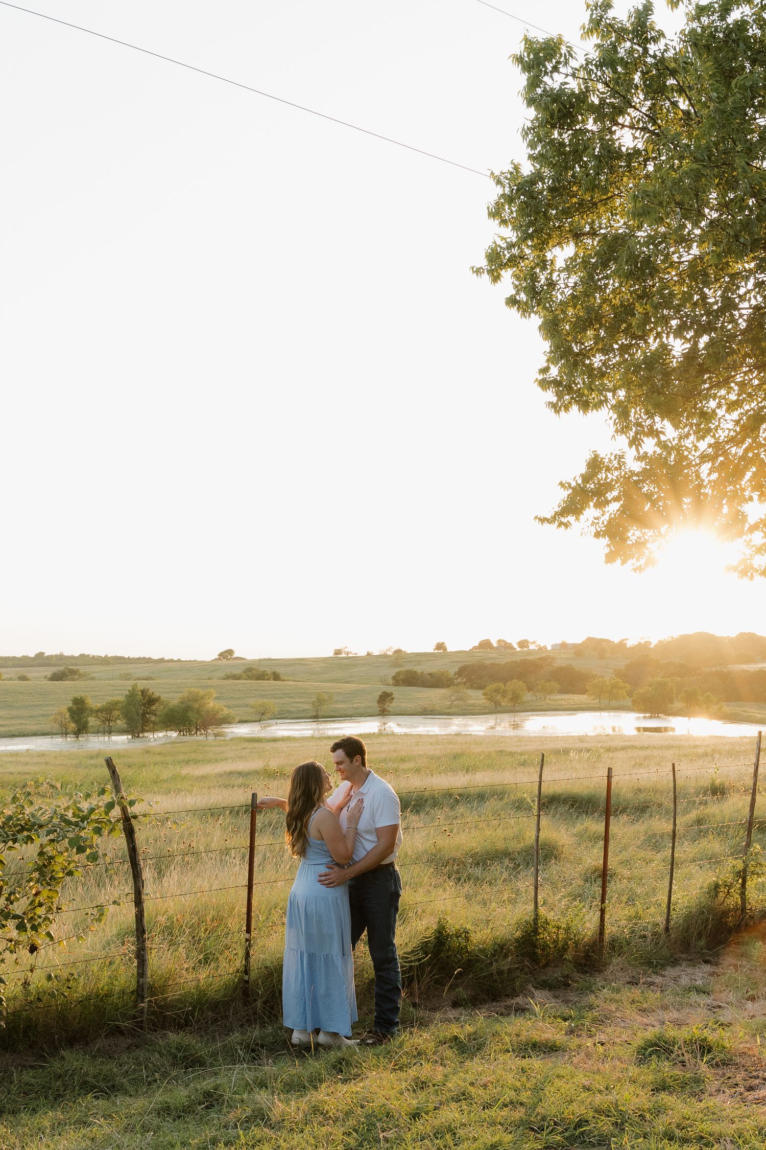 Couple holding each other close near a fence at sunset with a glowing pond in the background.