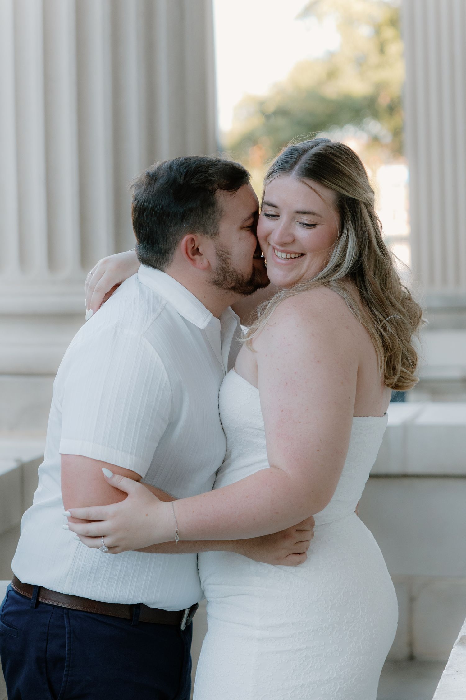 Couple embracing and laughing in white outfits during engagement photos by classical columns.
