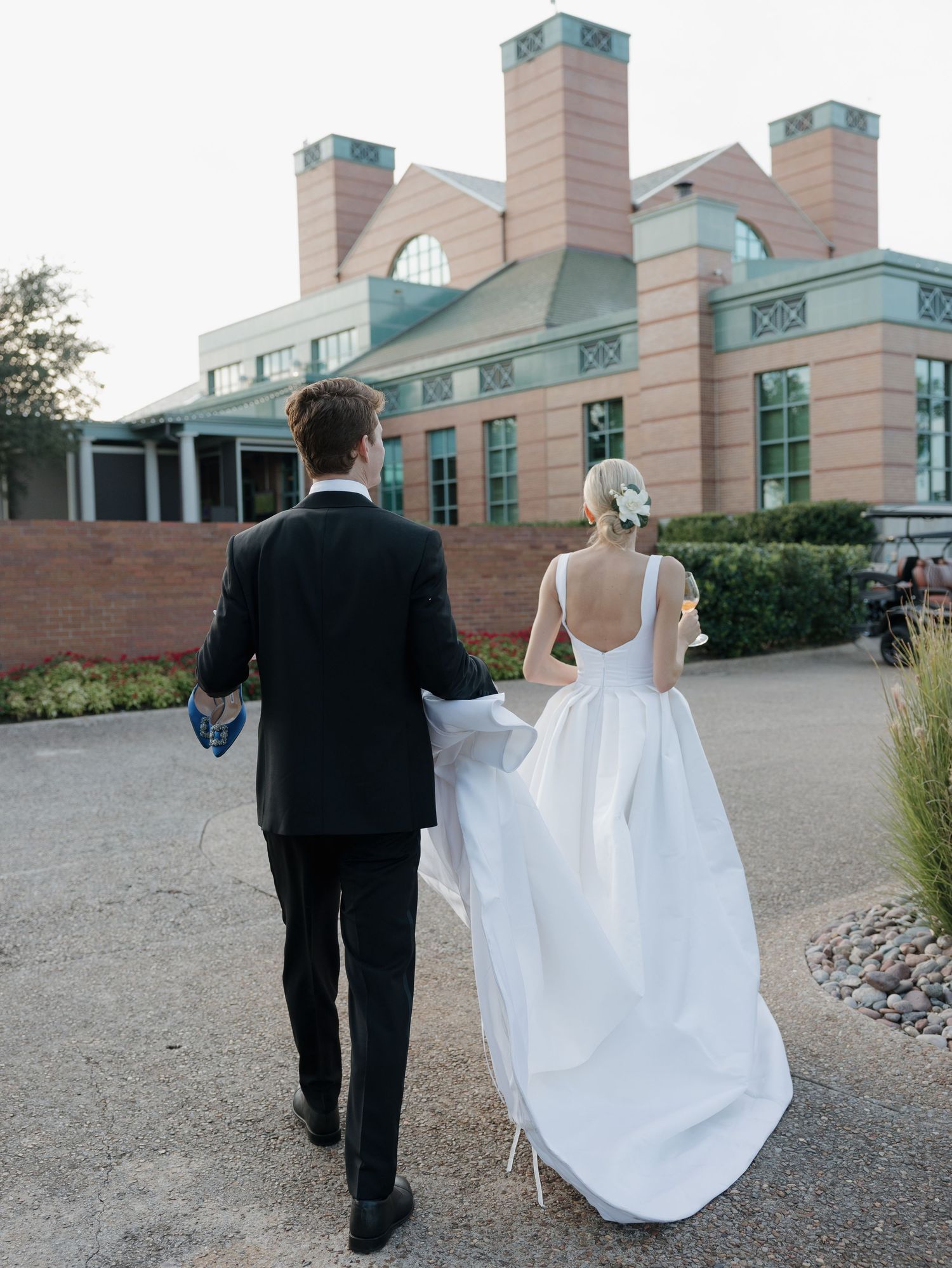 Bride and groom walking hand-in-hand toward elegant brick wedding venue, bride in backless white gown with train.