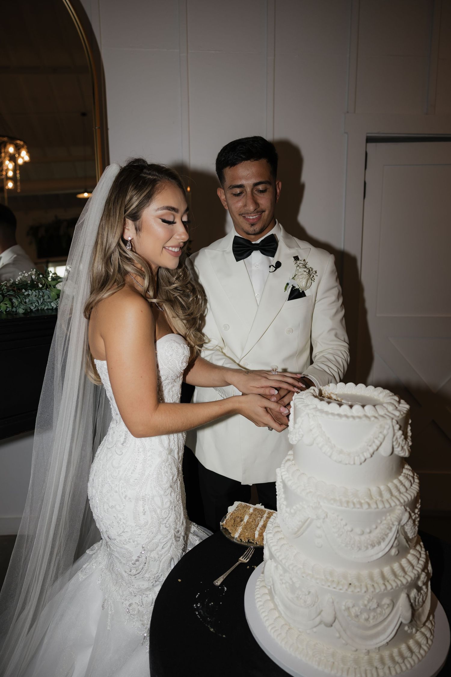 Bride and groom cut elegant white tiered wedding cake together at their indoor wedding reception.