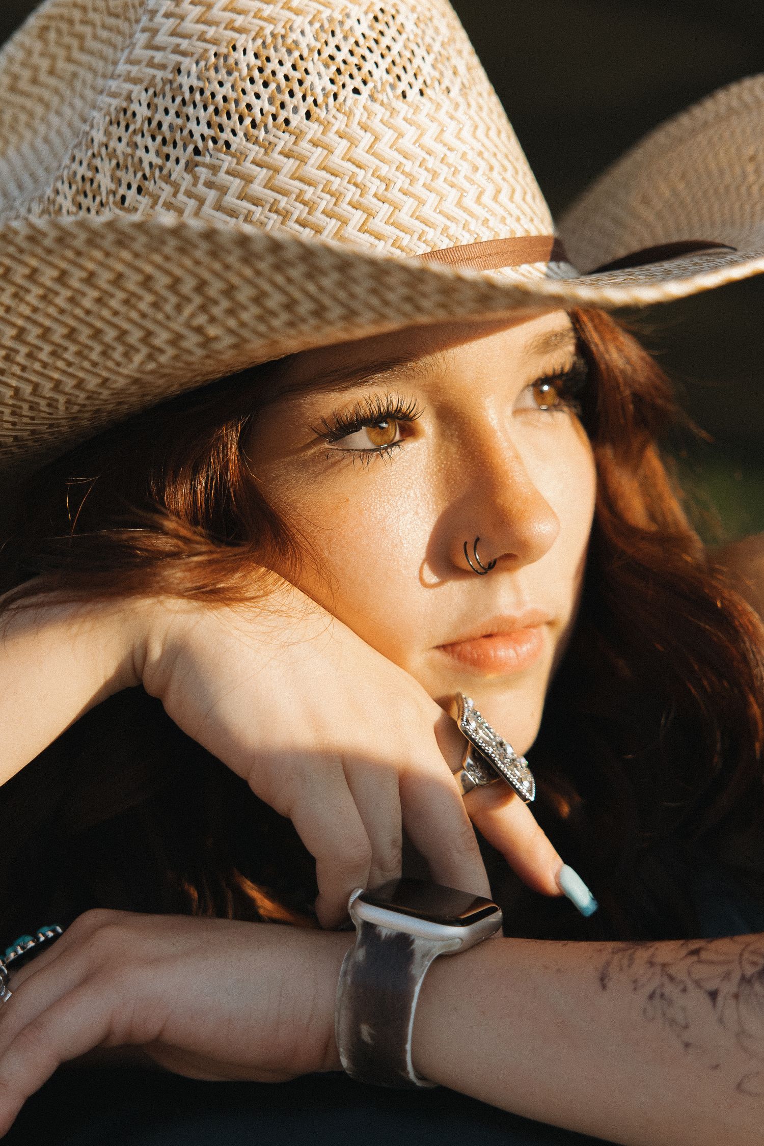Young woman in woven cowboy hat with silver rings and bracelet poses in warm golden sunlight.