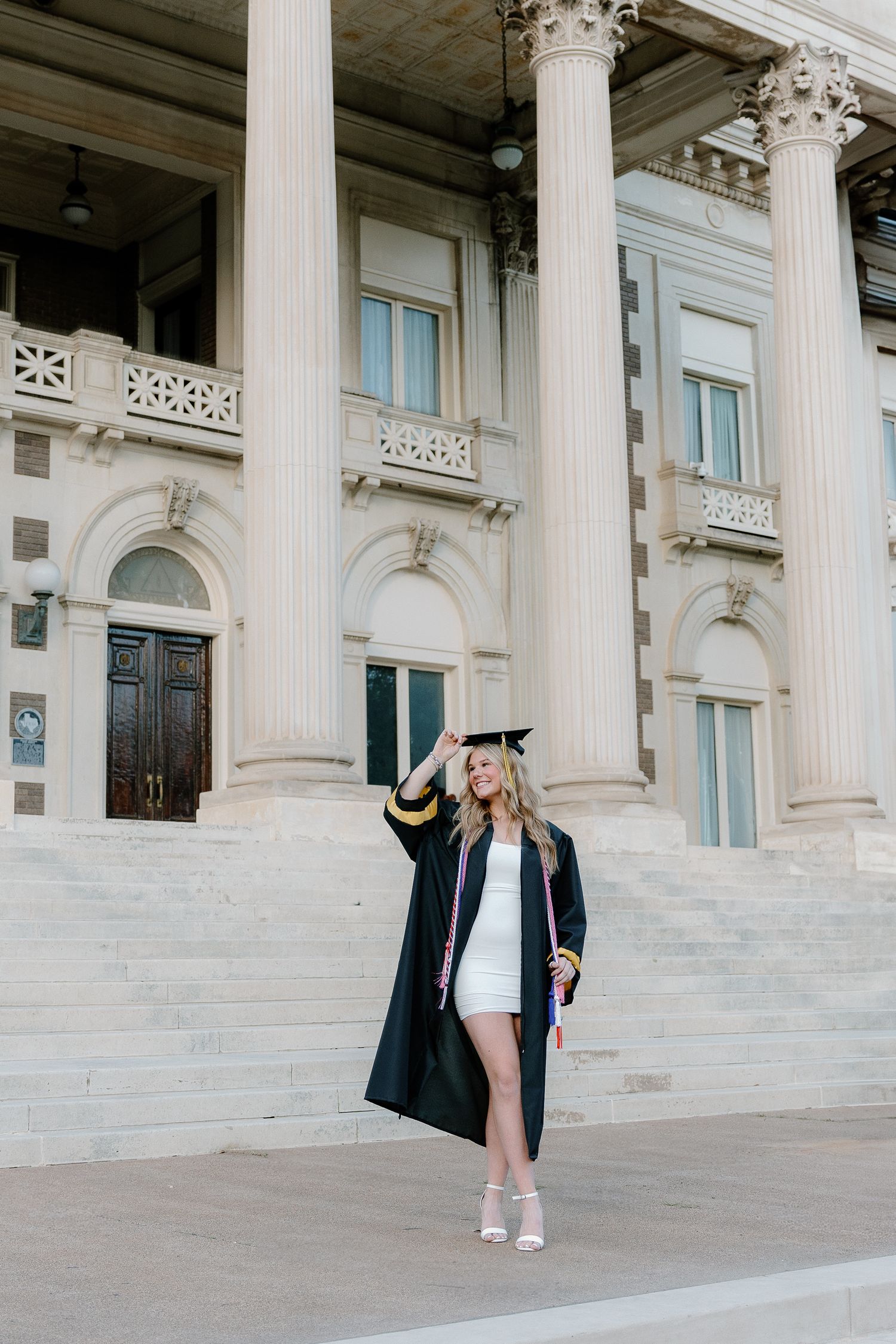 Blonde graduate in black cap and gown posing on steps of a neoclassical building holding a diploma.