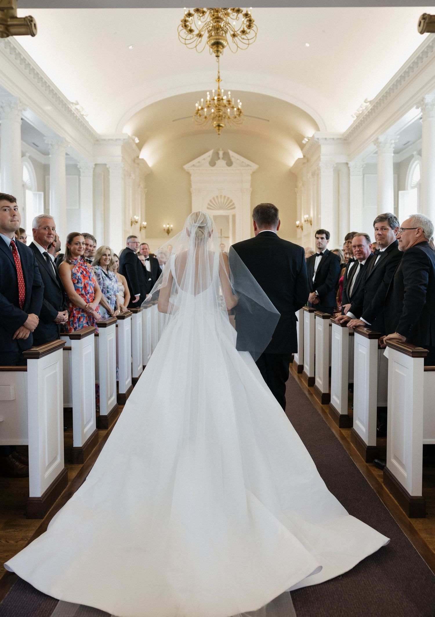 Bride in cathedral veil walking down elegant white chapel aisle escorted toward waiting groom.