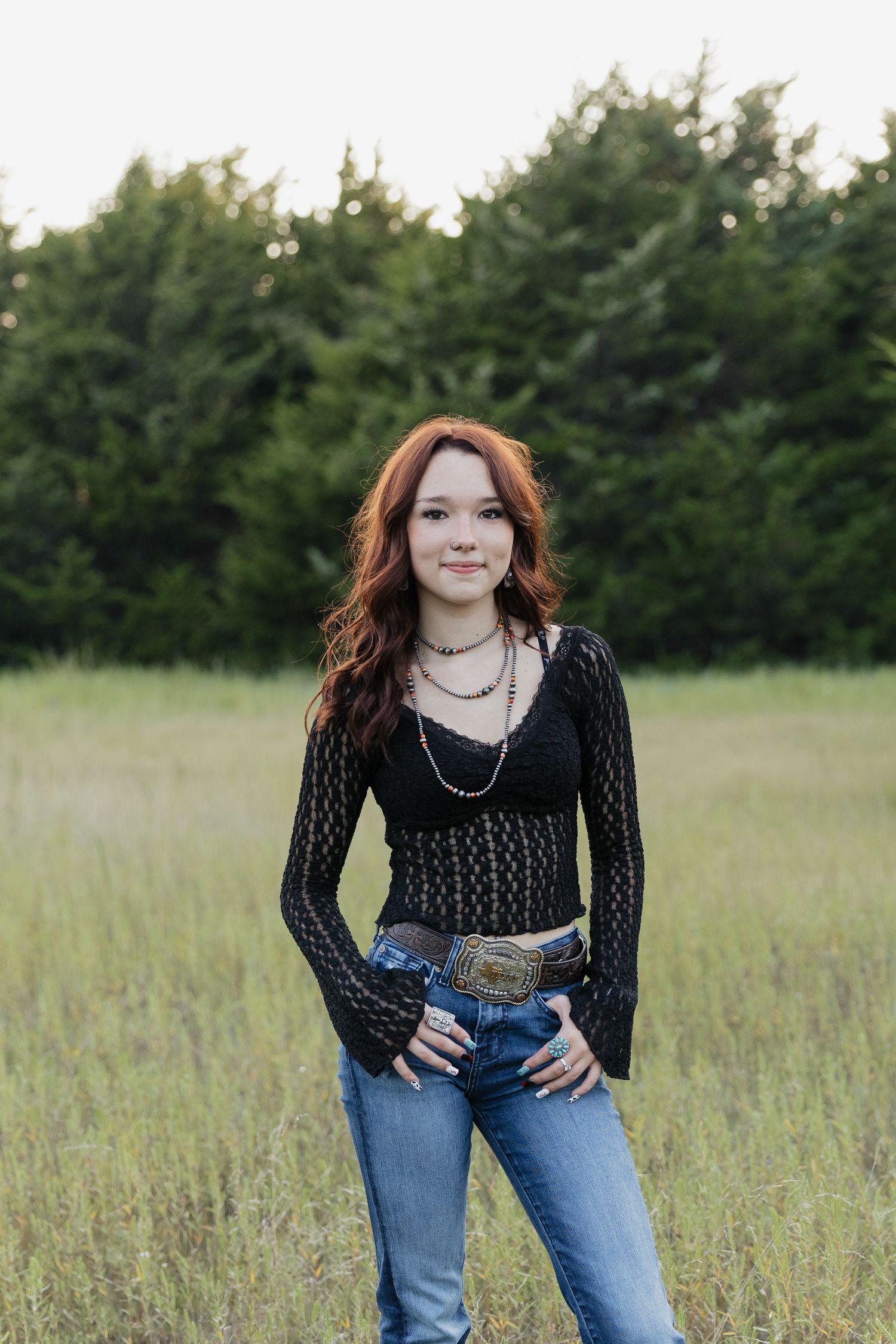 Young woman with red hair in a black lace top, jeans, and western belt buckle standing in a grassy field.