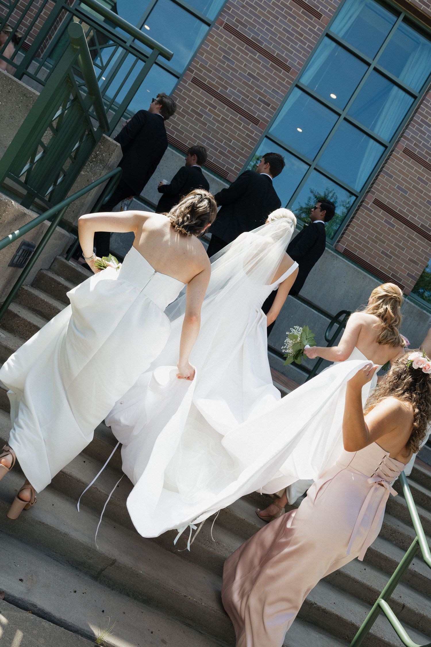 Bridesmaids in white and pink dresses helping bride carry her gown train up building steps outdoors.