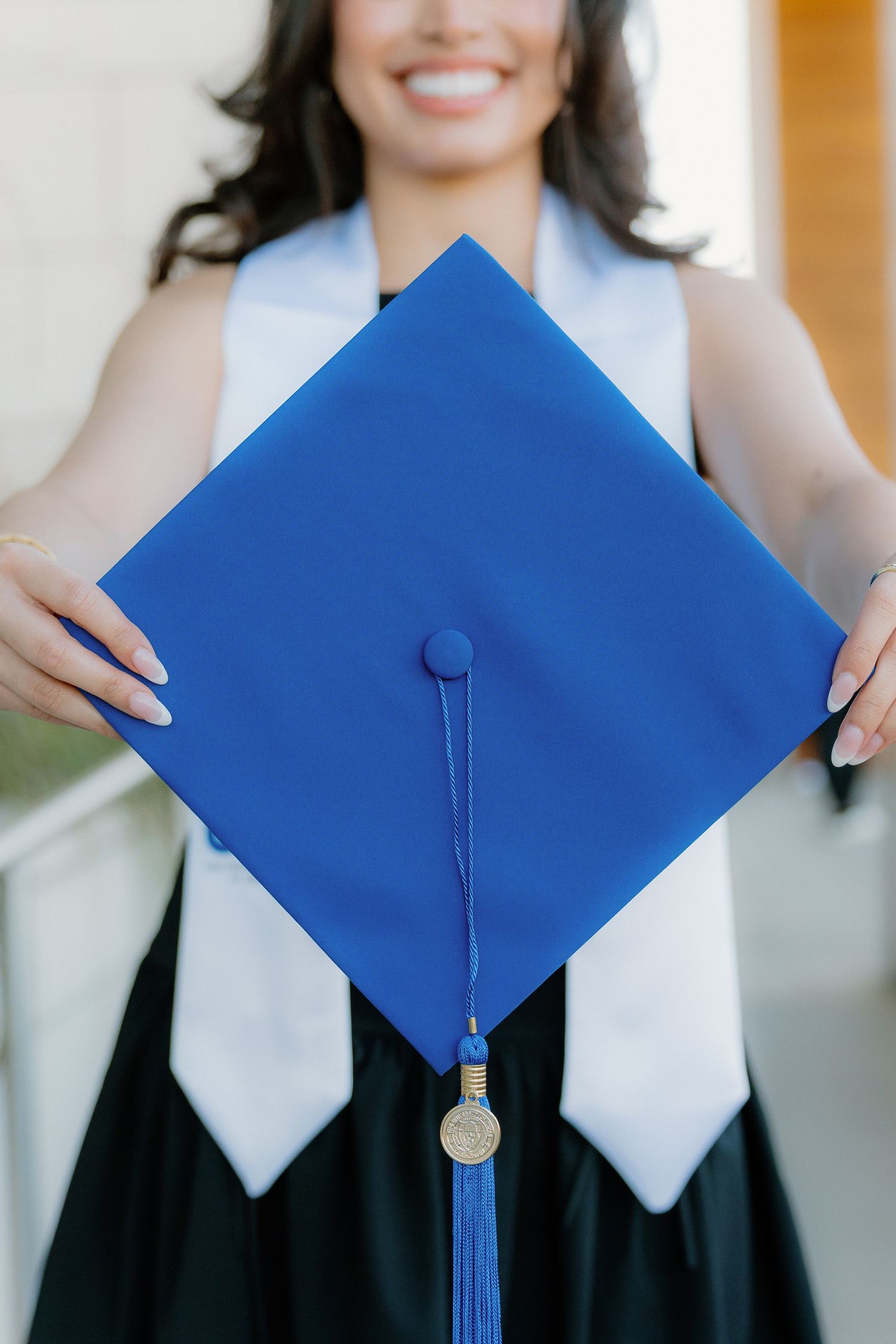 Graduate holding blue UTA mortarboard cap with gold tassel while wearing white graduation stole and black dress.