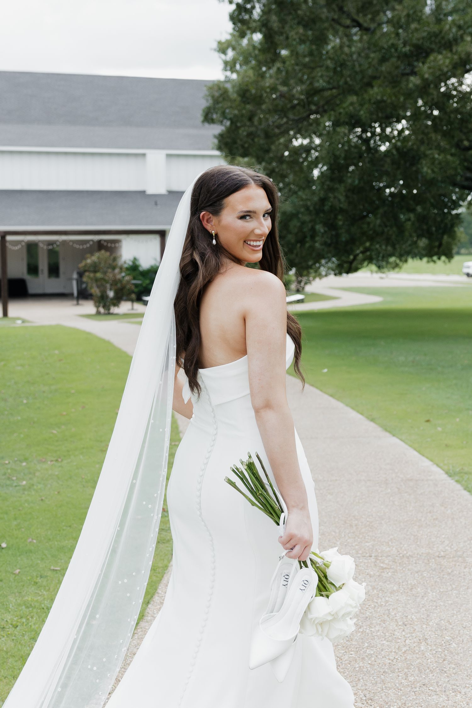 Elegant bride in fitted strapless gown and cathedral veil holds white bouquet on garden pathway.