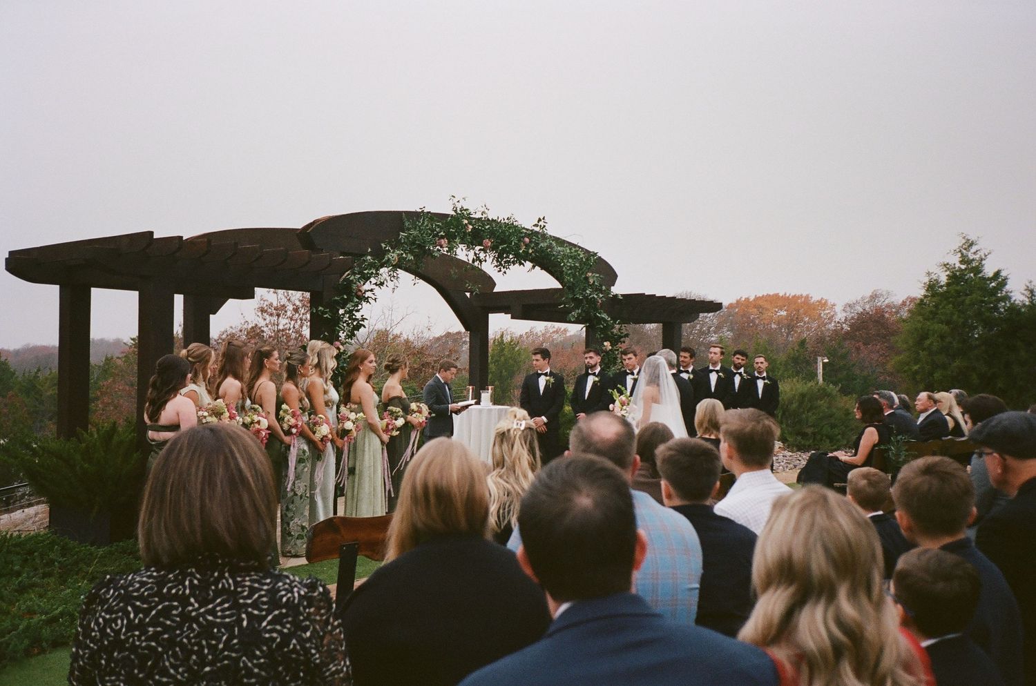 Outdoor wedding ceremony with bridal party under circular greenery arch on overcast day.