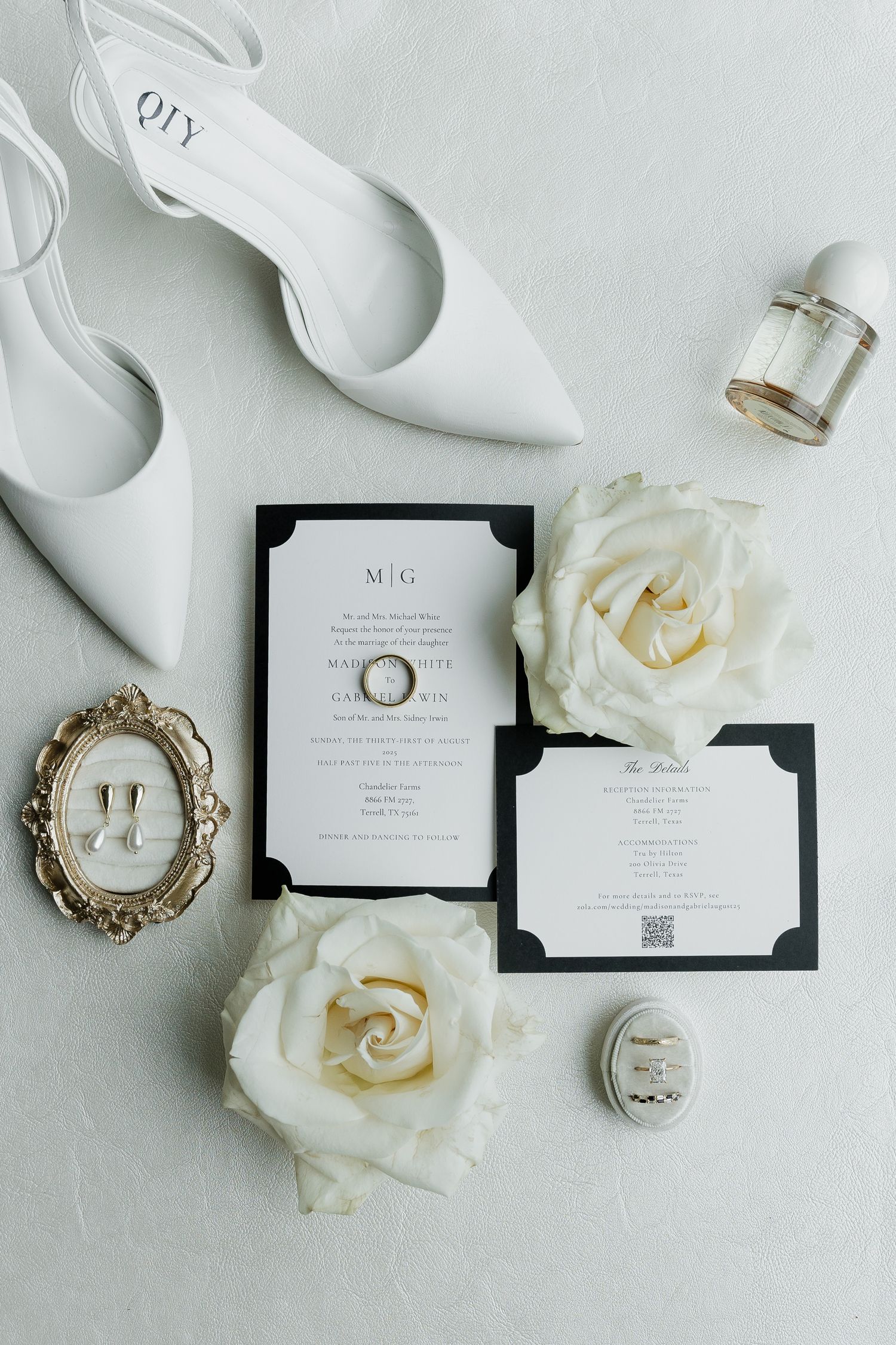 Black and white wedding invitation flatlay with white roses, heels, perfume bottle, and monogram details on white background.