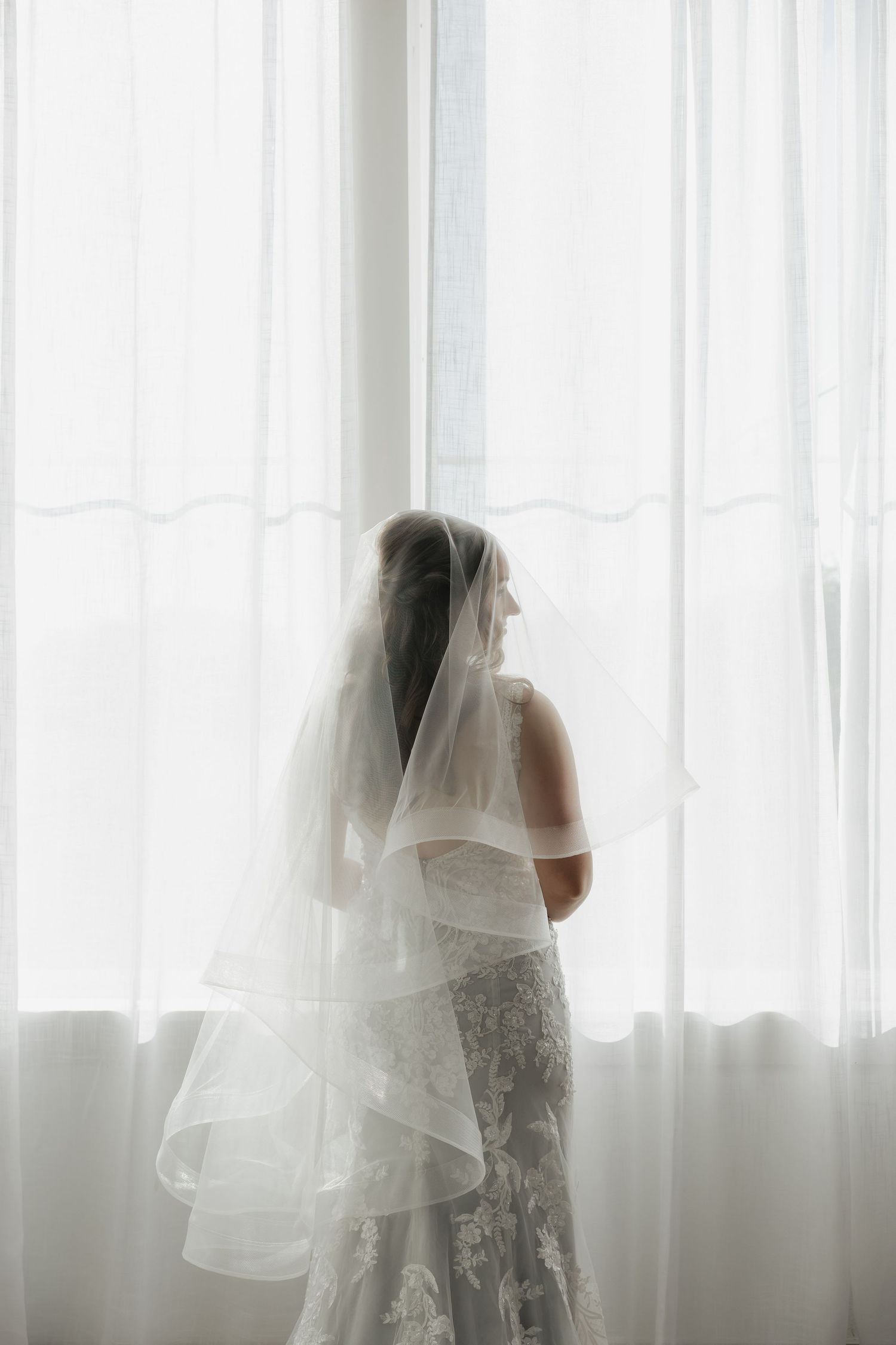 Bride in lace wedding gown and layered veil standing by bright window.