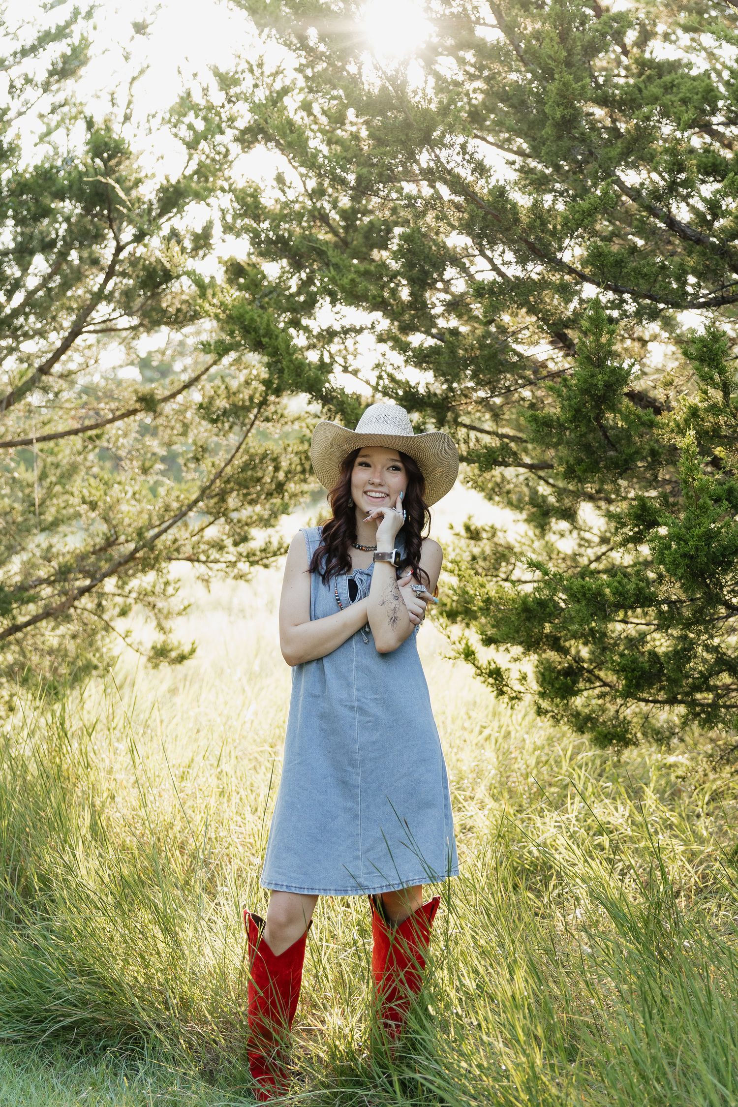 Young woman in denim dress, red cowboy boots, and straw hat standing in a sunlit grassy field near trees.