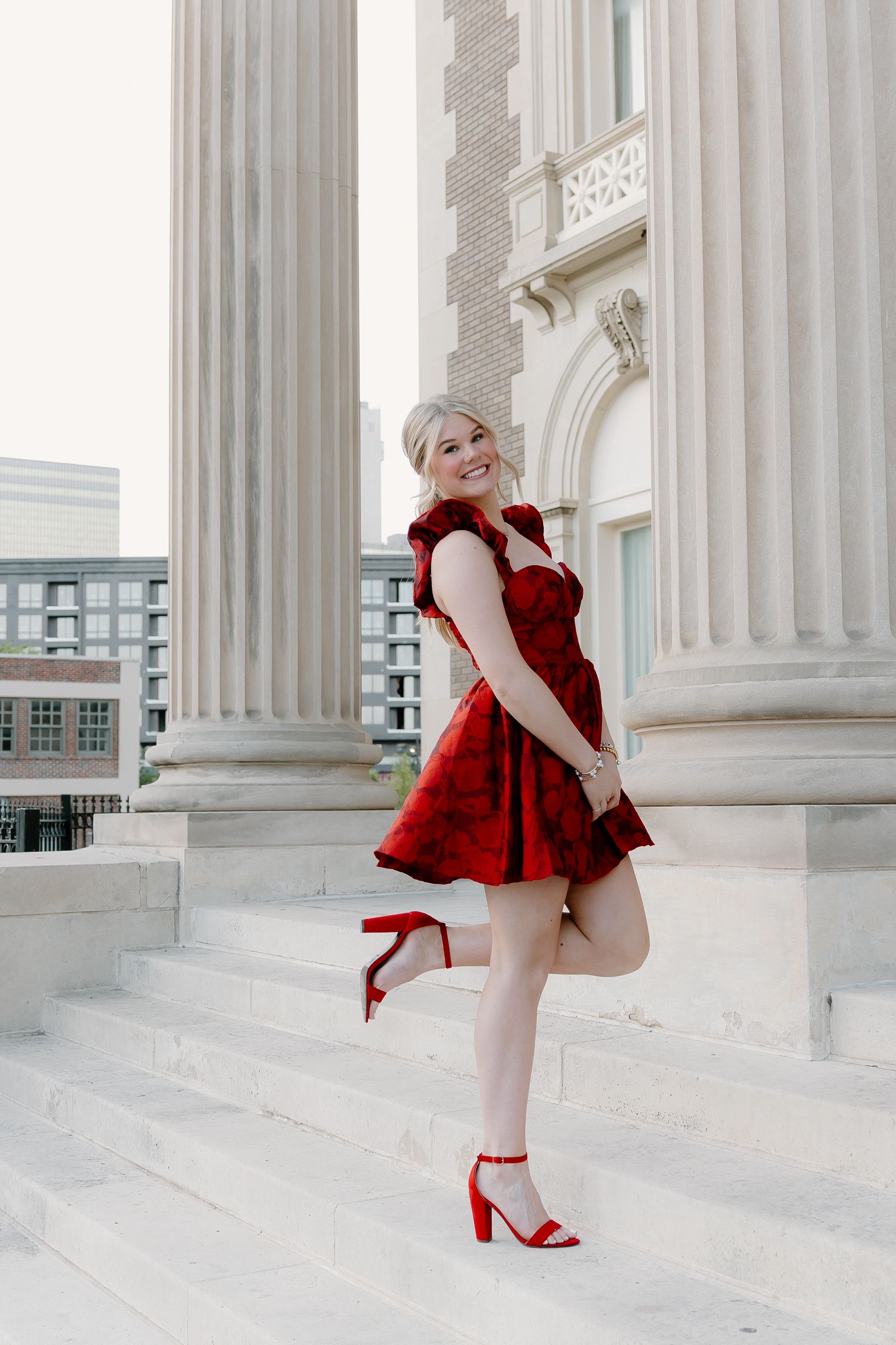 Young woman in red floral fit-and-flare dress with red block heels posing playfully on marble building steps.