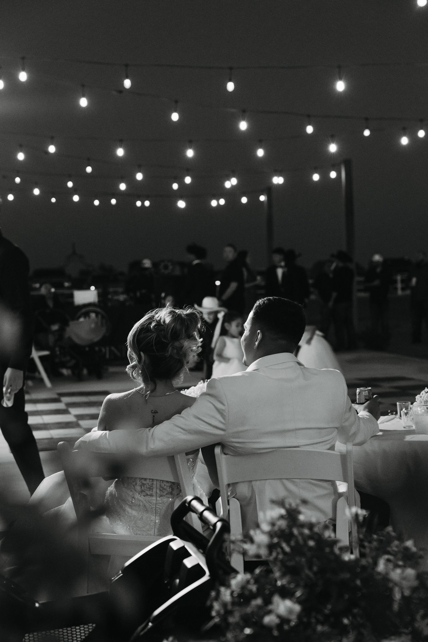 Black and white photo of bride and groom seated at reception under glowing string lights.