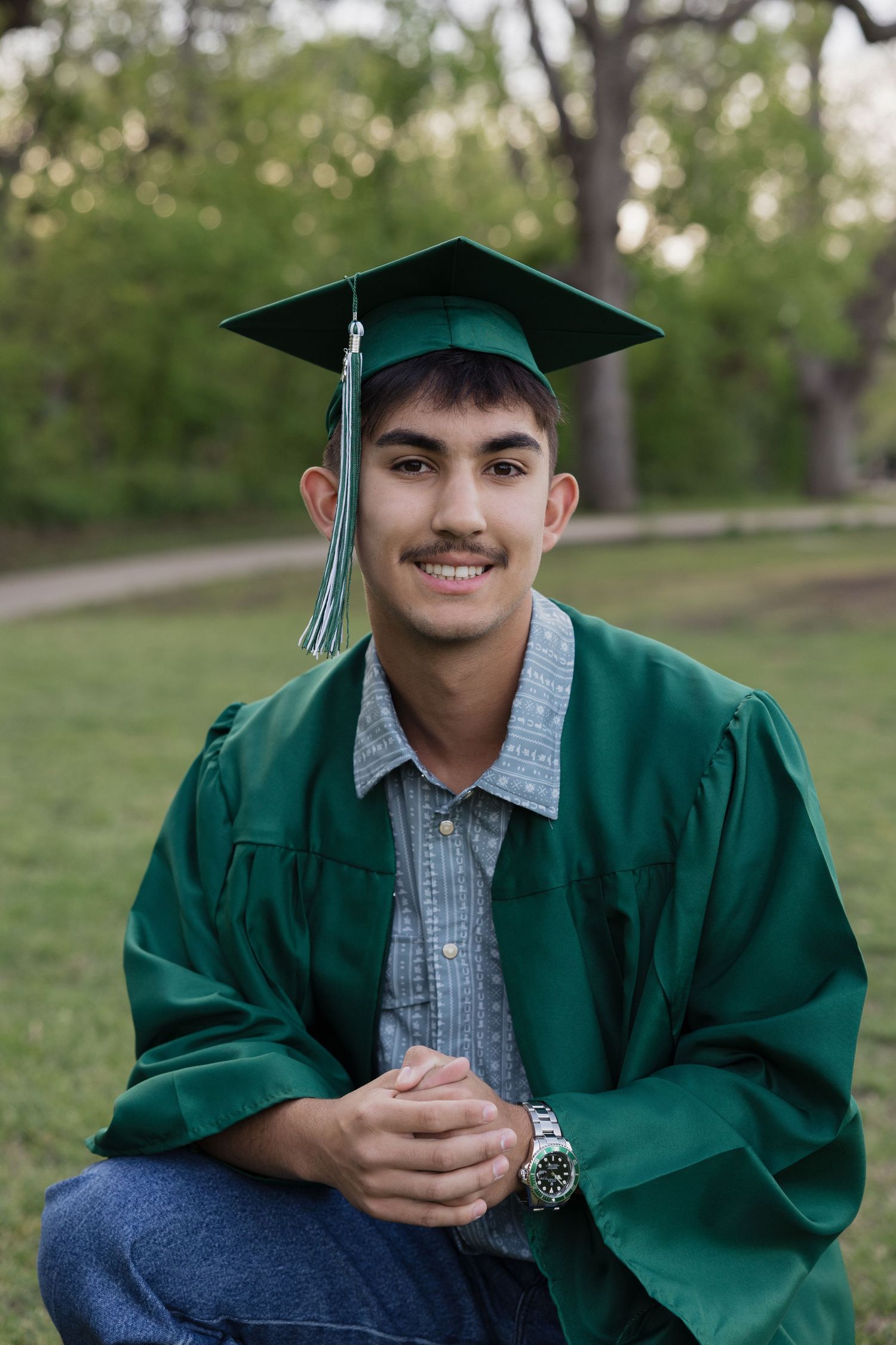 Young man smiling in green graduation cap and gown, seated outdoors in a park setting.