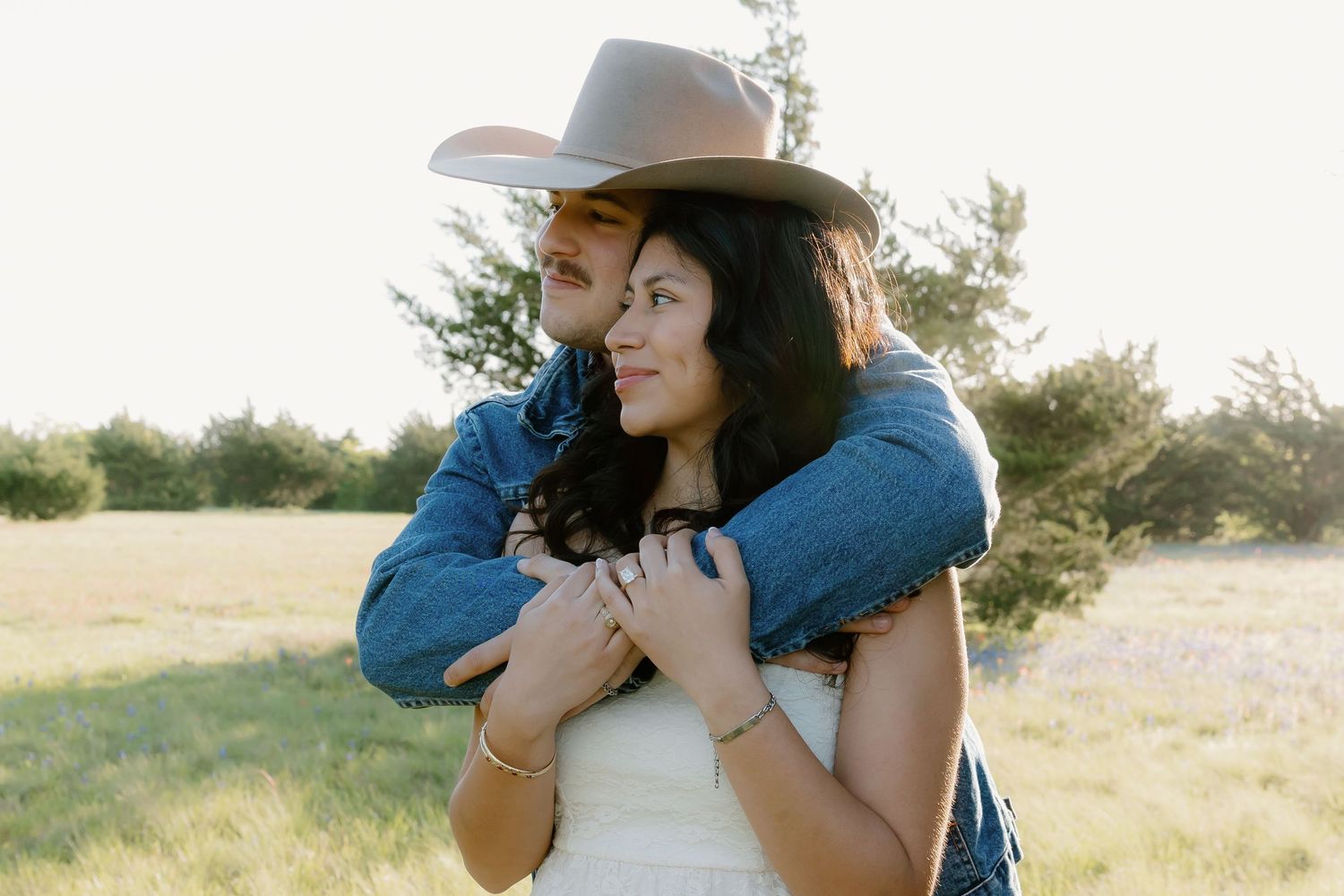 Woman in cowboy hat and white dress embraced from behind by partner in sunny meadow.