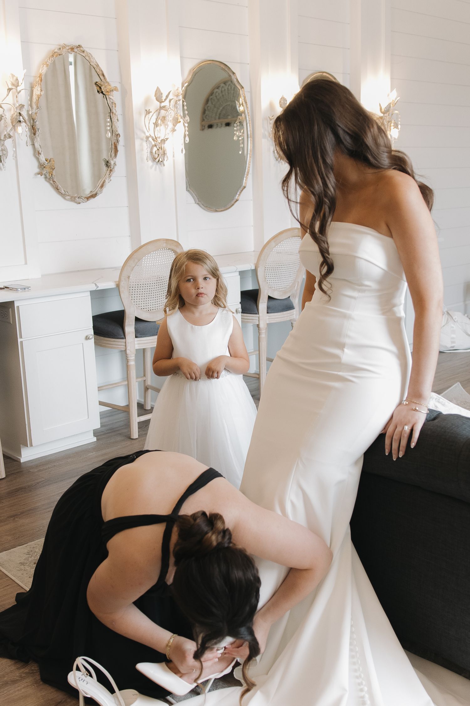 Bridesmaid adjusts brides shoes while flower girl watches in elegant white bridal suite with oval mirrors.