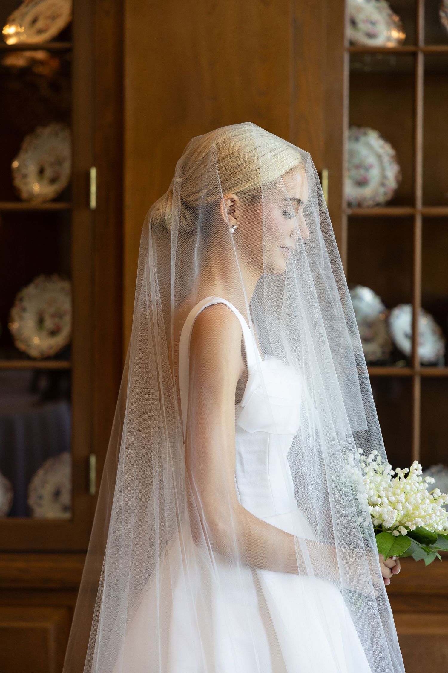 Elegant bride in white gown and cathedral veil holding lily of the valley bouquet standing by wooden cabinet.