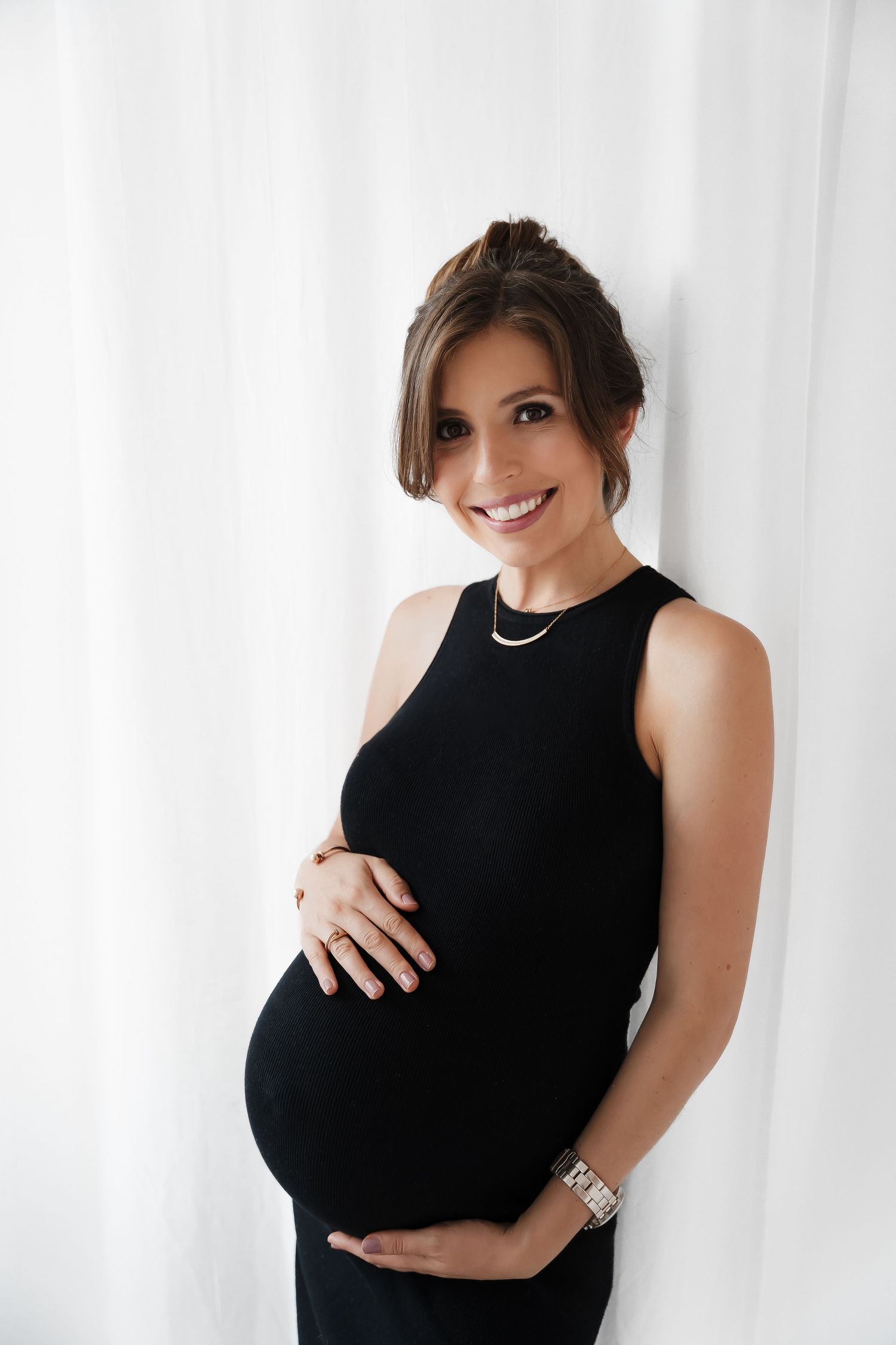 Maternity portrait with woman in black sleeveless dress against white curtain.