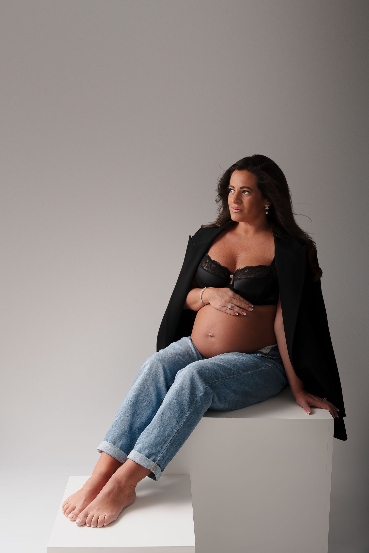 Artistic maternity photo of an expectant mother in jeans and black top sitting casually on a white cube.