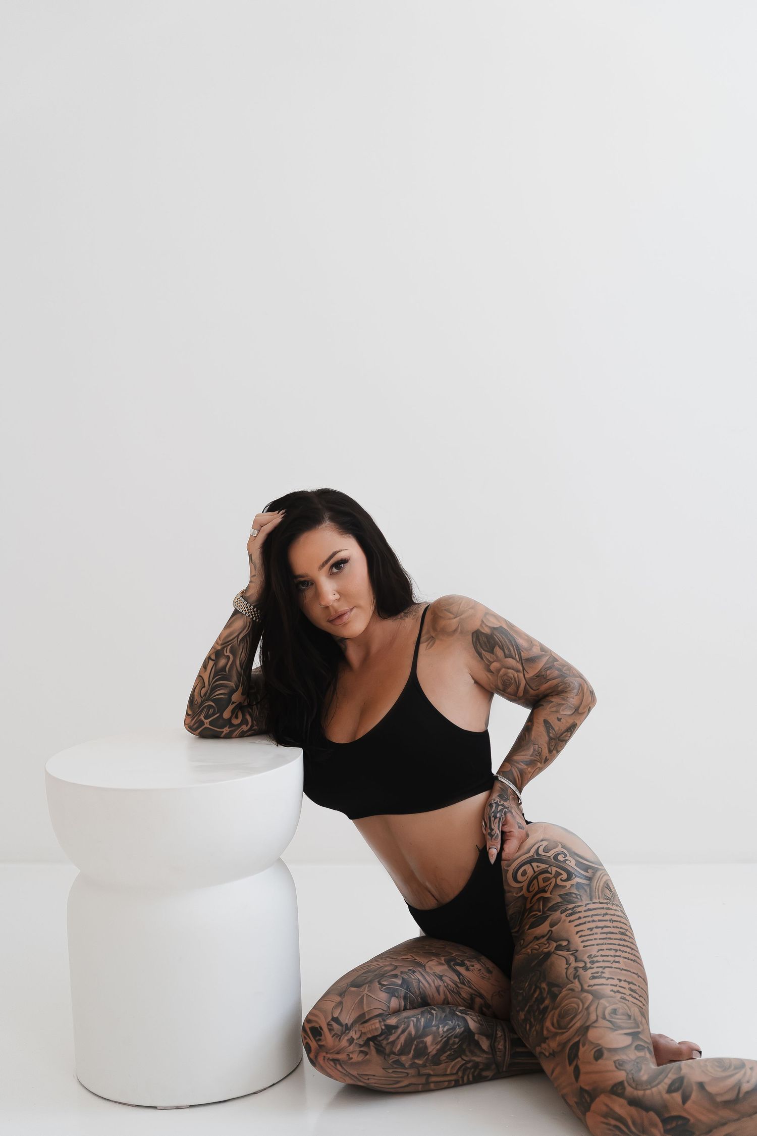 Tattooed model in black athletic wear poses with white cylindrical prop against white background.