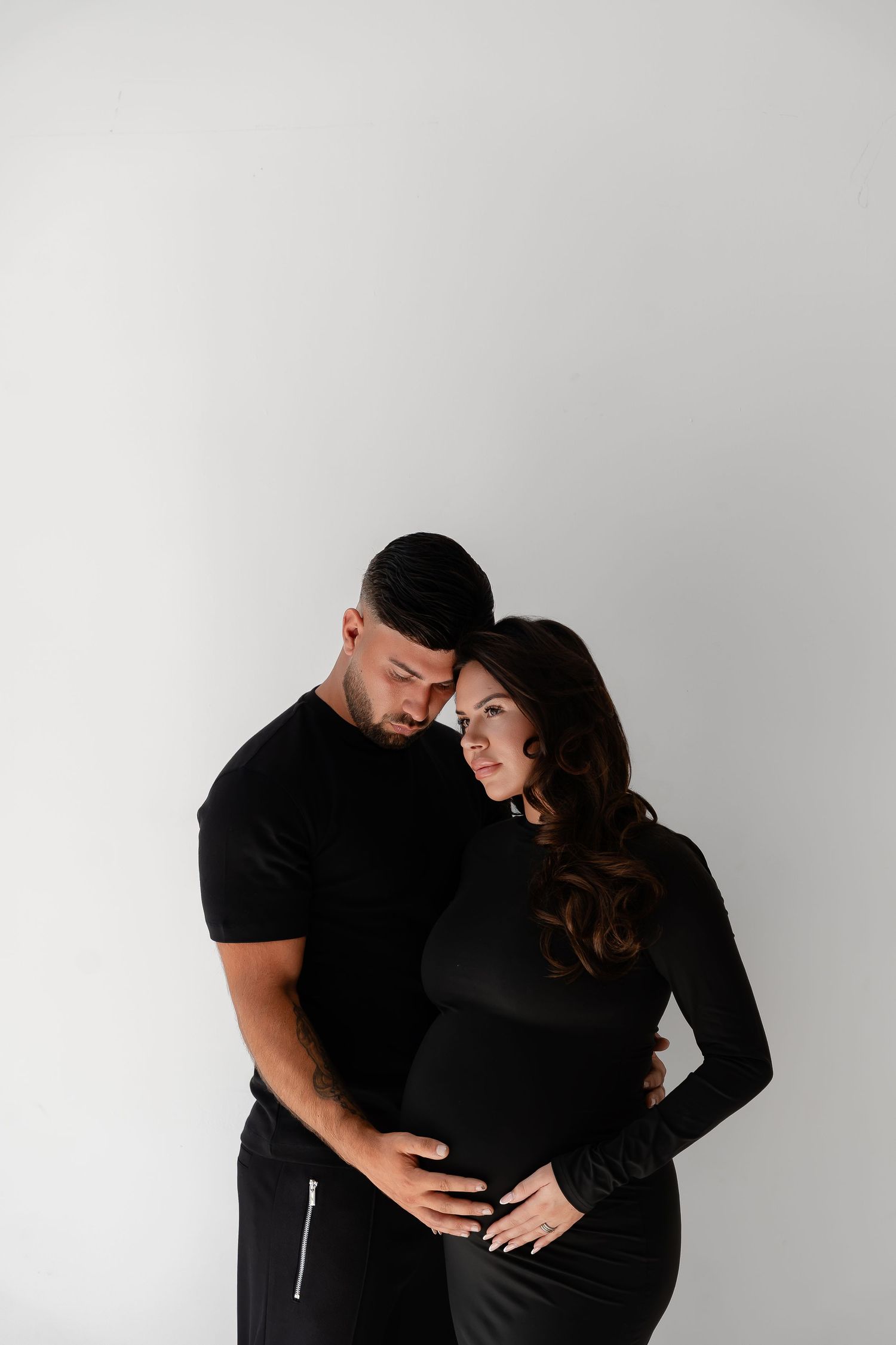 A couple in matching black outfits tenderly embrace, the woman visibly pregnant, against a white studio background.