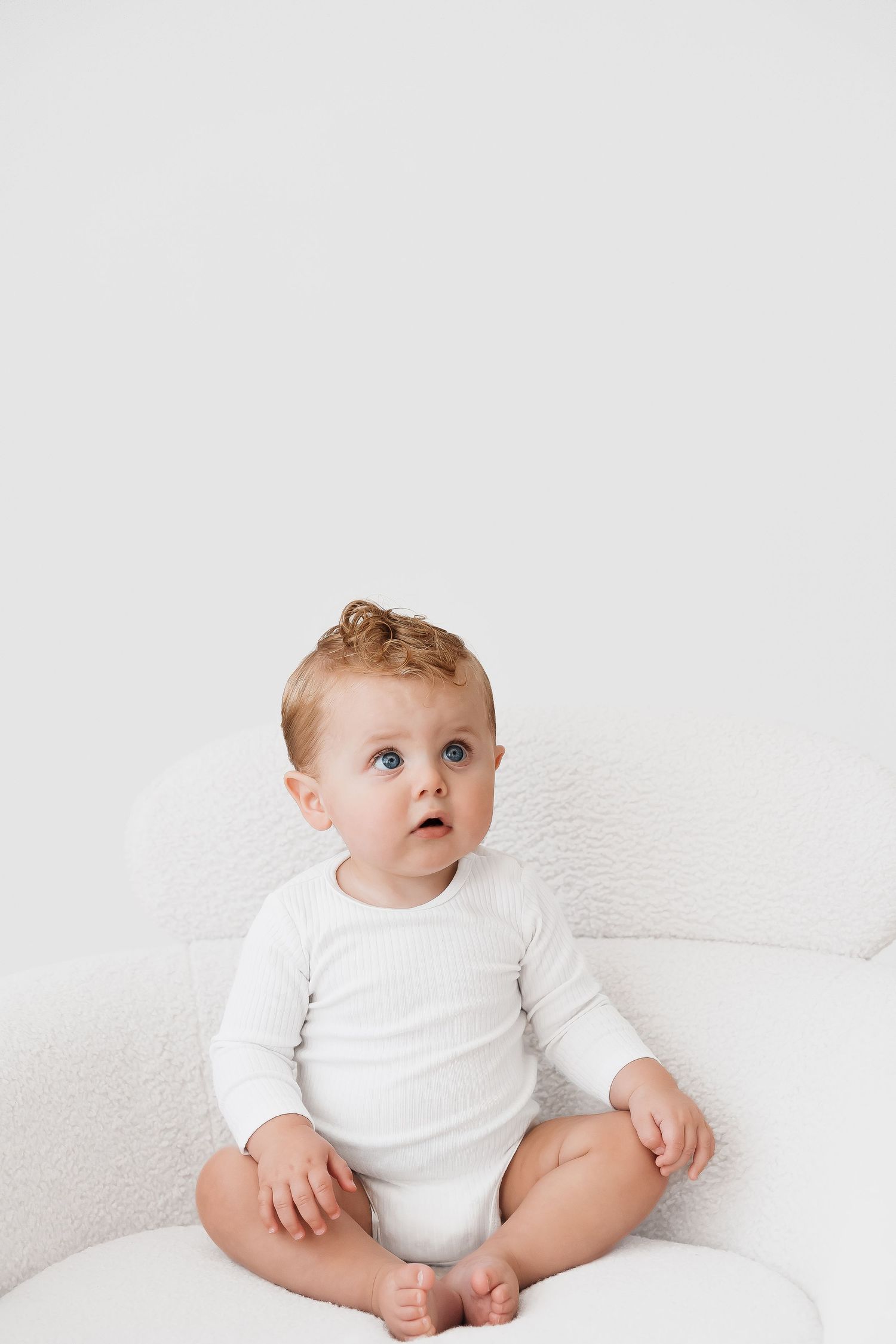 Baby in white onesie sitting on white couch during minimalist photo shoot.