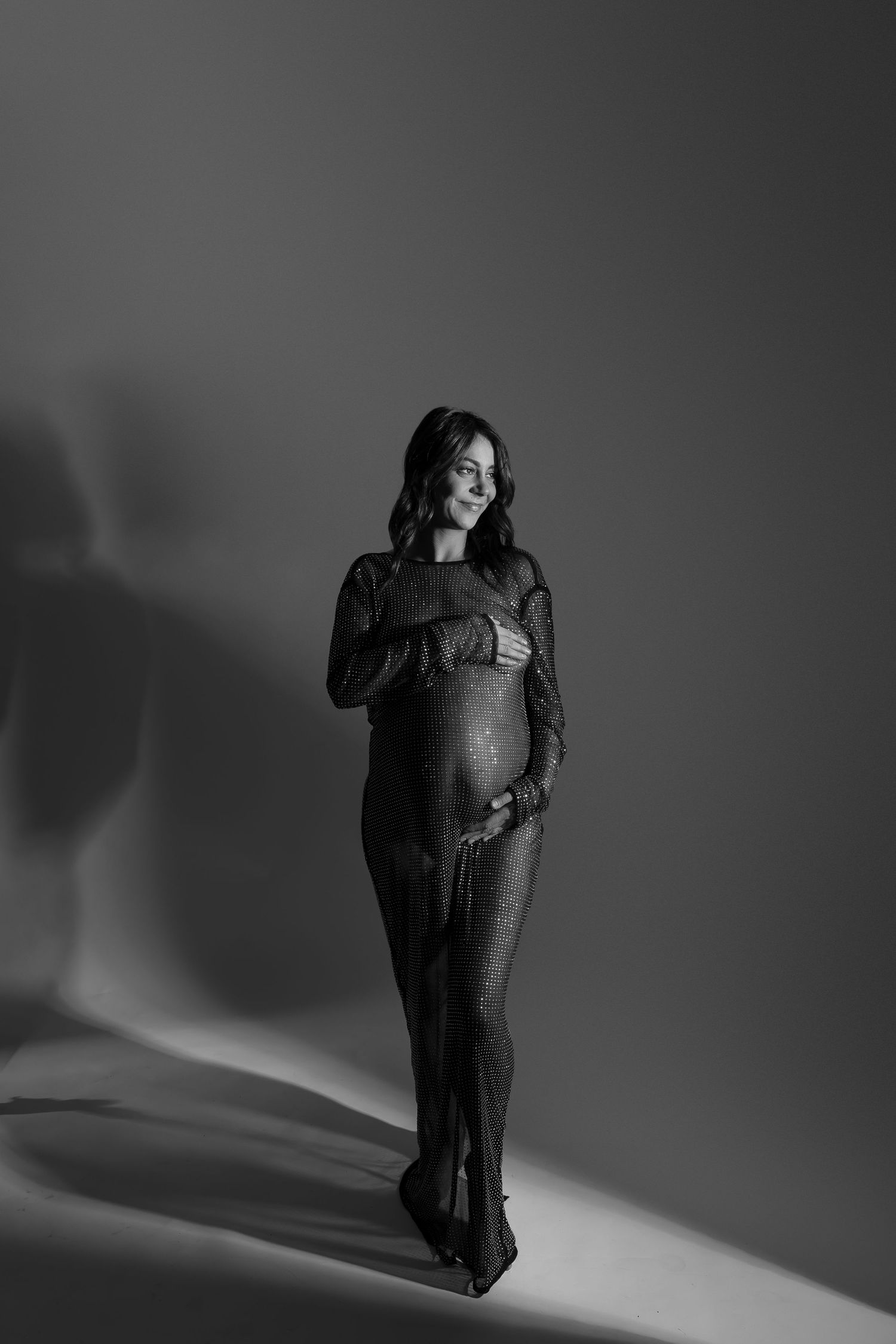 Artistic black and white maternity photo showing an expectant mother in a dark dress against a shadowy background.