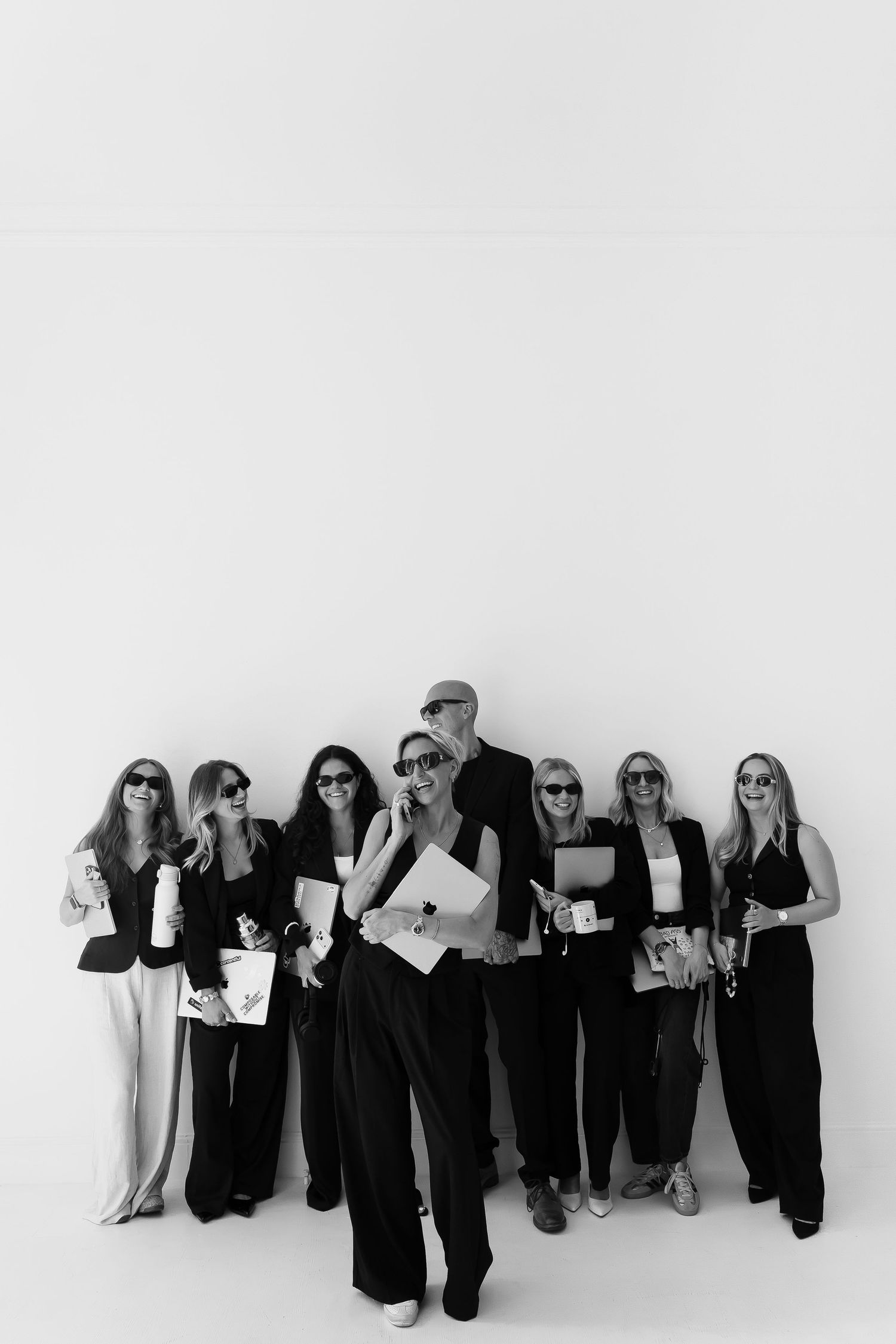 Black and white photo of a stylish group of professionals in sunglasses and suits posing confidently against a white wall.