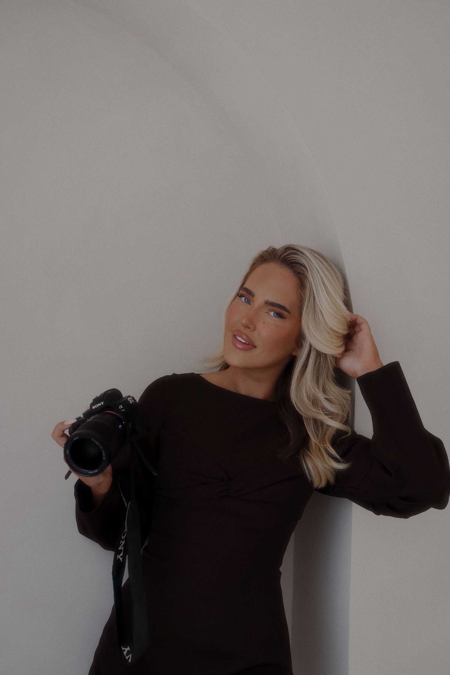 Professional photographer in black sweater poses with camera against neutral background.