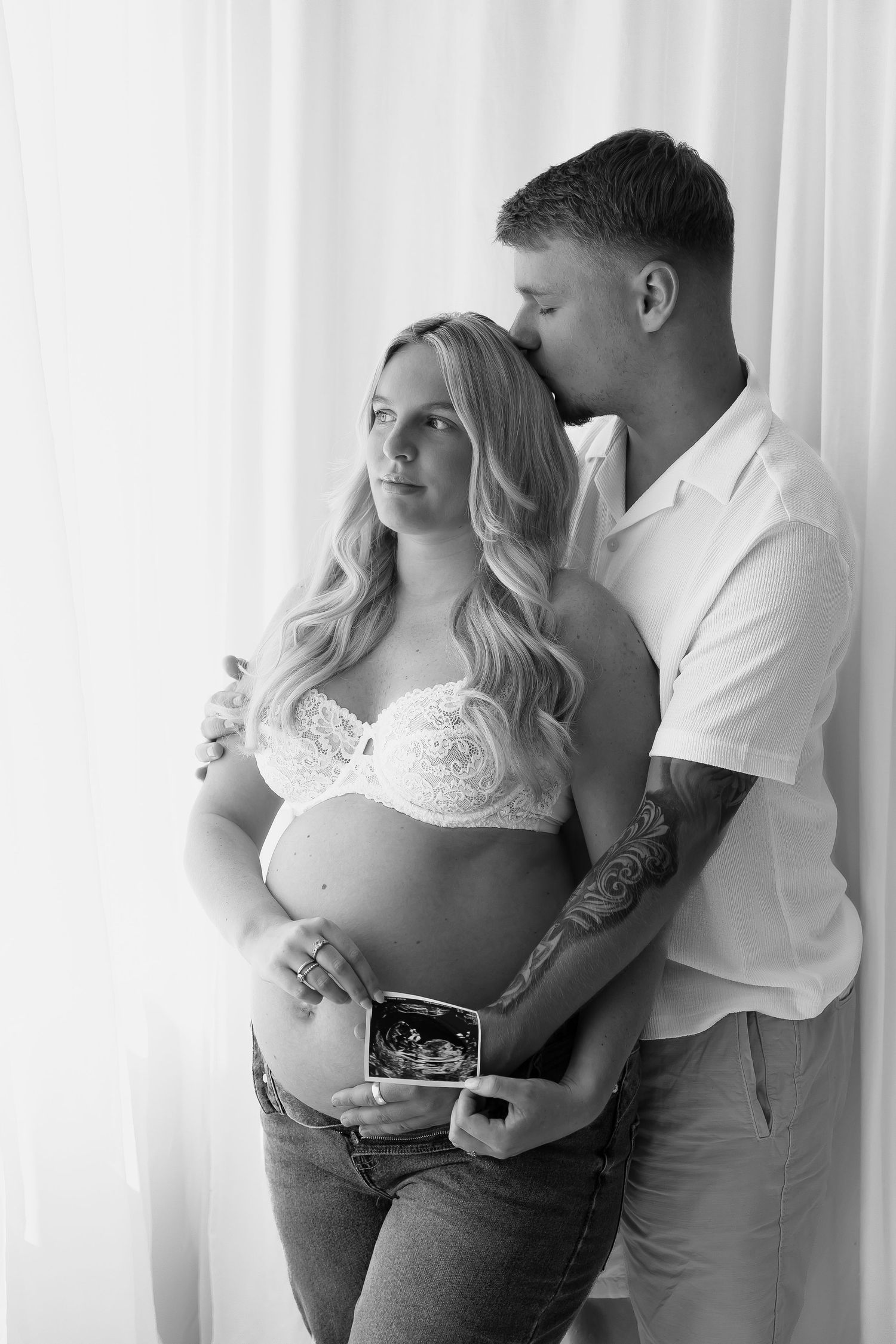 Intimate maternity photoshoot featuring couple embracing while holding ultrasound photo against white curtains.