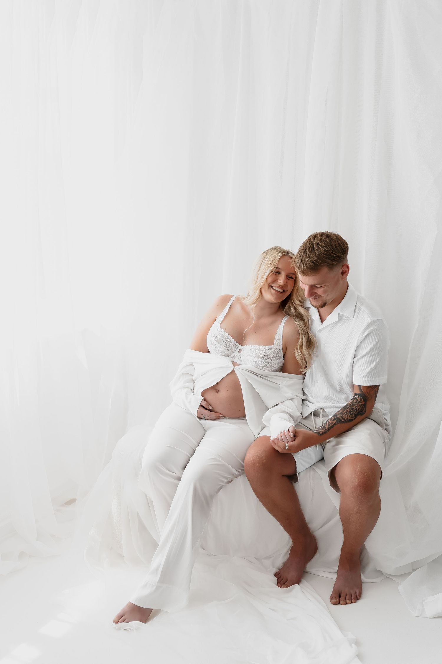 Couple in white clothing poses intimately on floor during maternity photoshoot.
