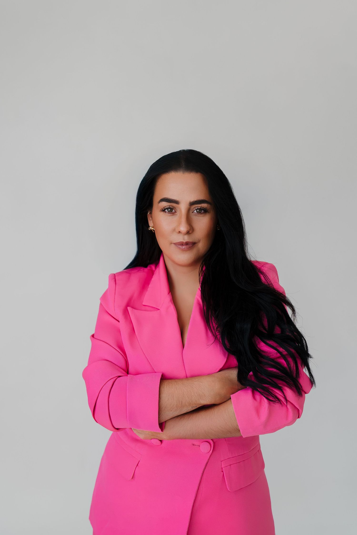Woman in bright pink blazer with arms crossed, dark hair, confident pose against light gray background.