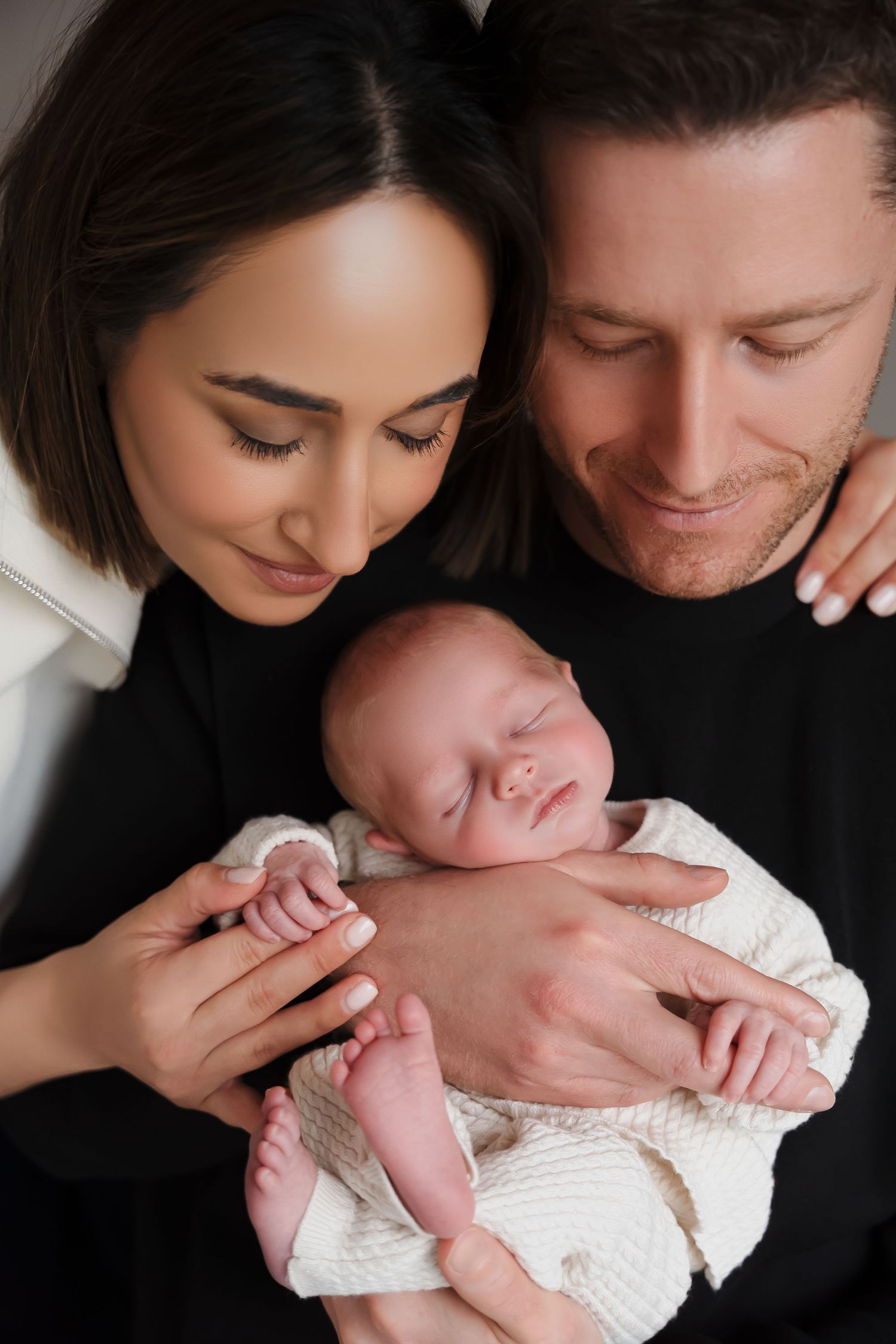 Happy parents in black outfits tenderly holding their sleeping newborn baby wrapped in white blanket.