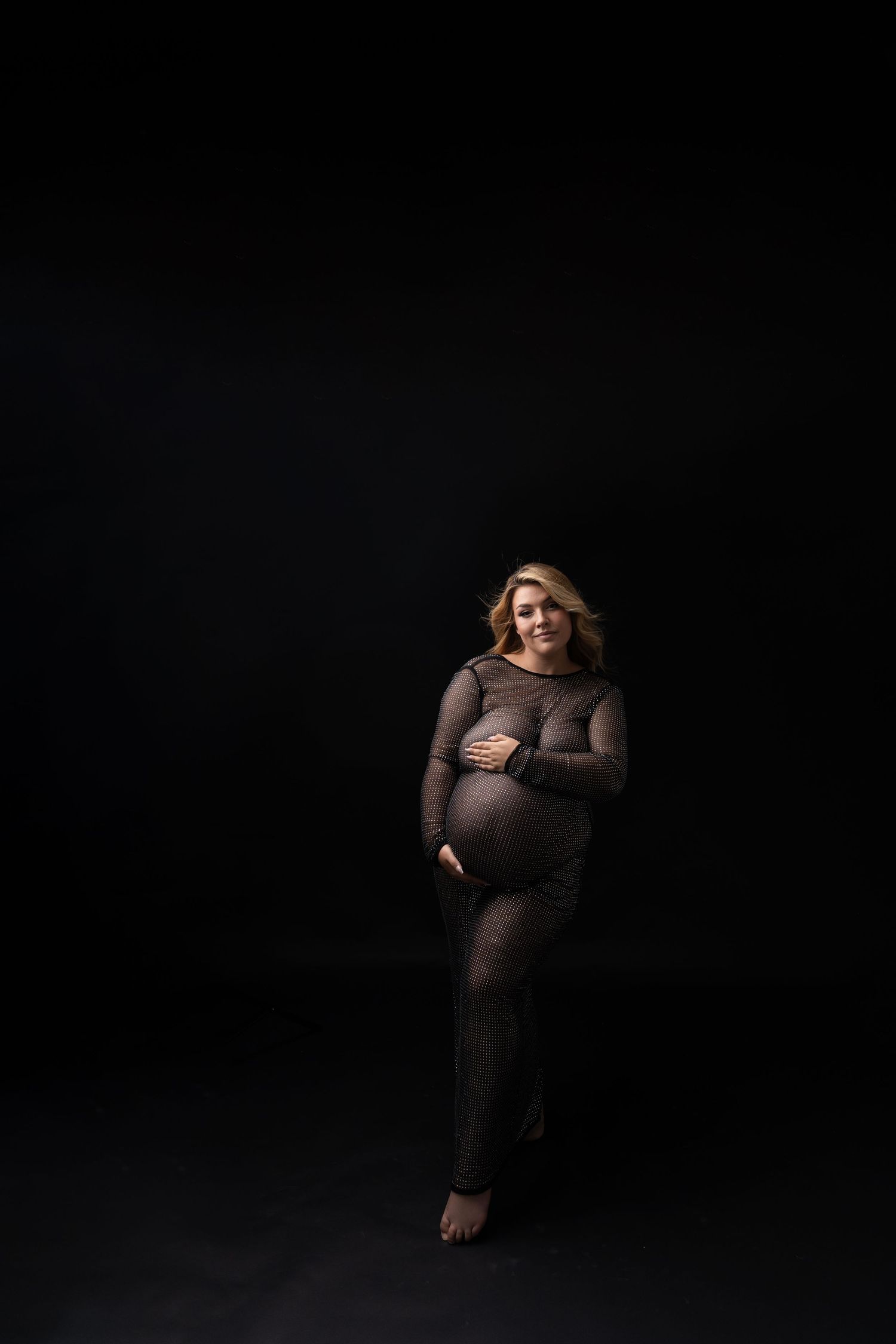 Dramatic low-key maternity portrait in sheer black outfit against dark background.