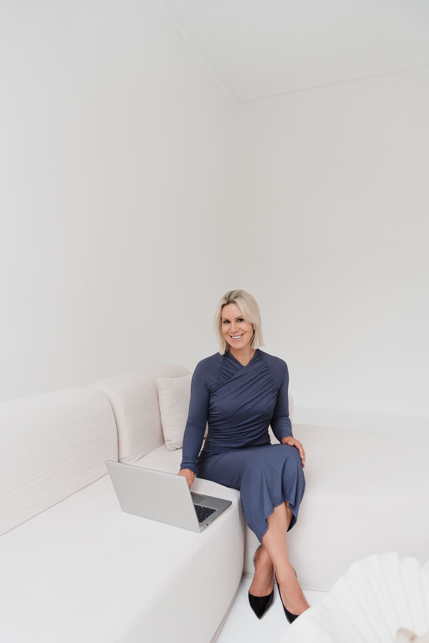Smiling blonde woman in blue dress sitting on white sofa with laptop in bright minimalist room.