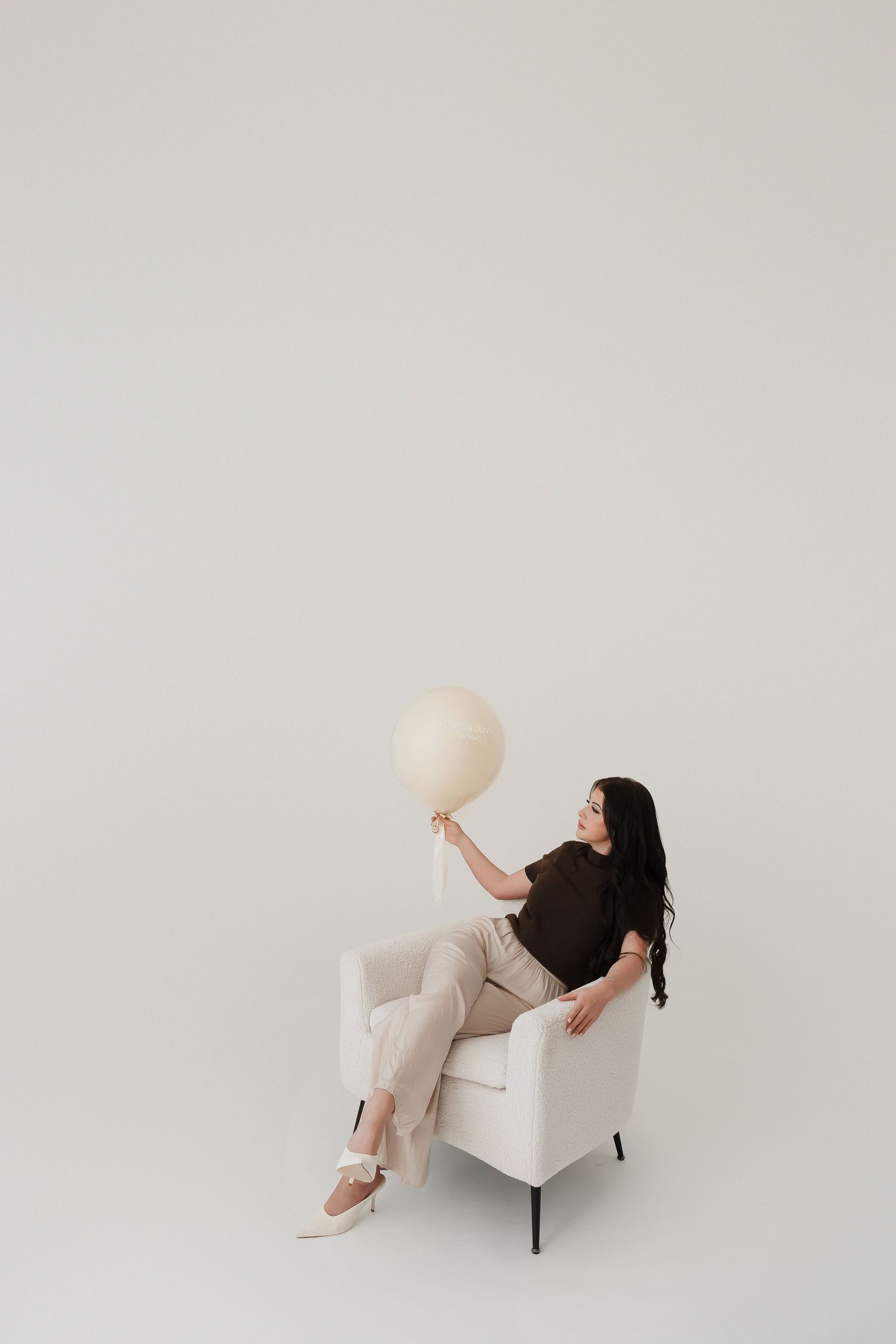 Woman in beige pants and brown top sitting in white chair holding a large cream balloon against light background.